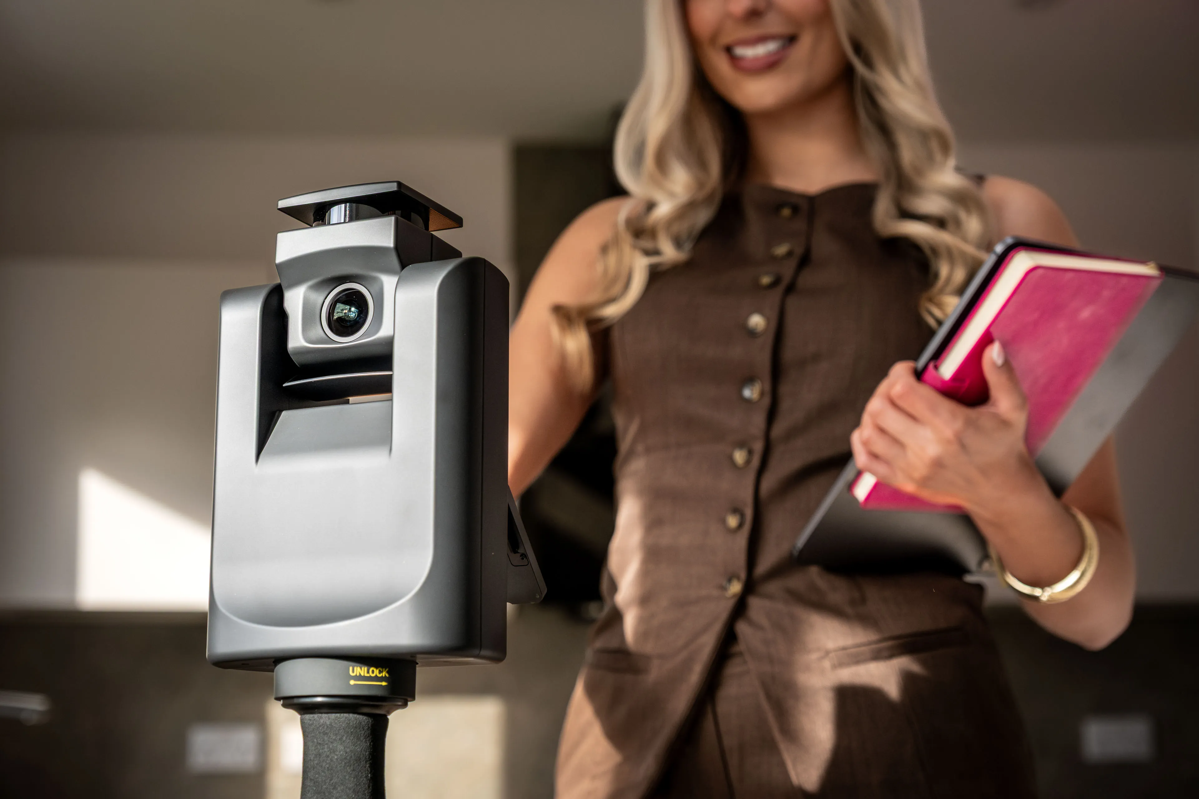 A woman in a professional outfit holding a pink notebook and tablet, smiling at a camera set on a tripod. The setting is bright, conveying a professional tone.