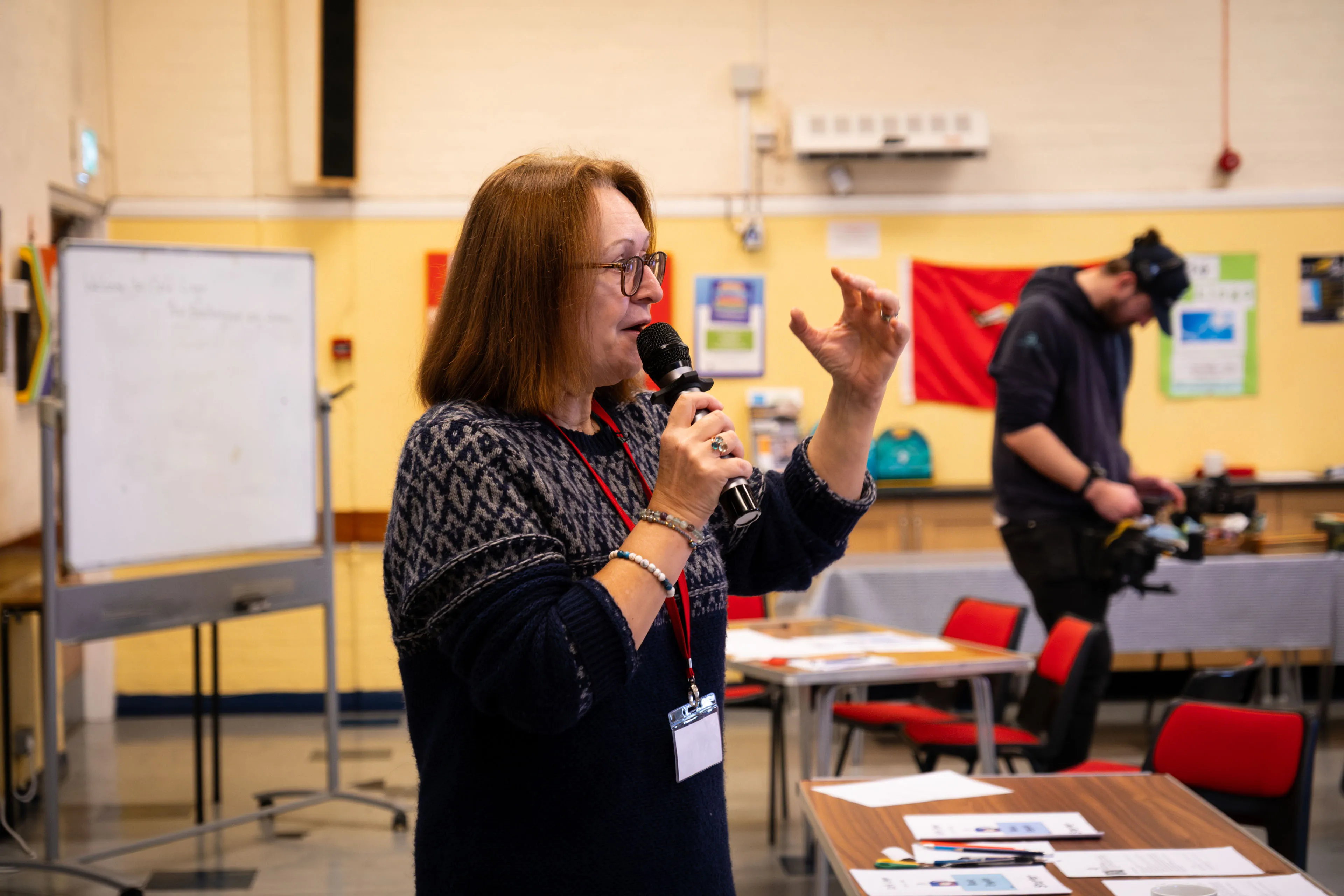 A woman with glasses speaks into a microphone, gesturing with one hand. She stands in a classroom with tables, chairs, and a whiteboard. The atmosphere is engaging and educational.