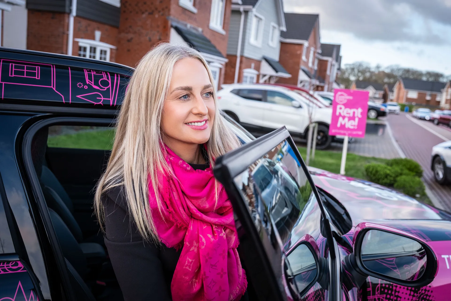 A woman with long blonde hair and a pink scarf smiles while stepping out of a colourful car in a suburban neighbourhood. A "Rent Me!" sign is visible.