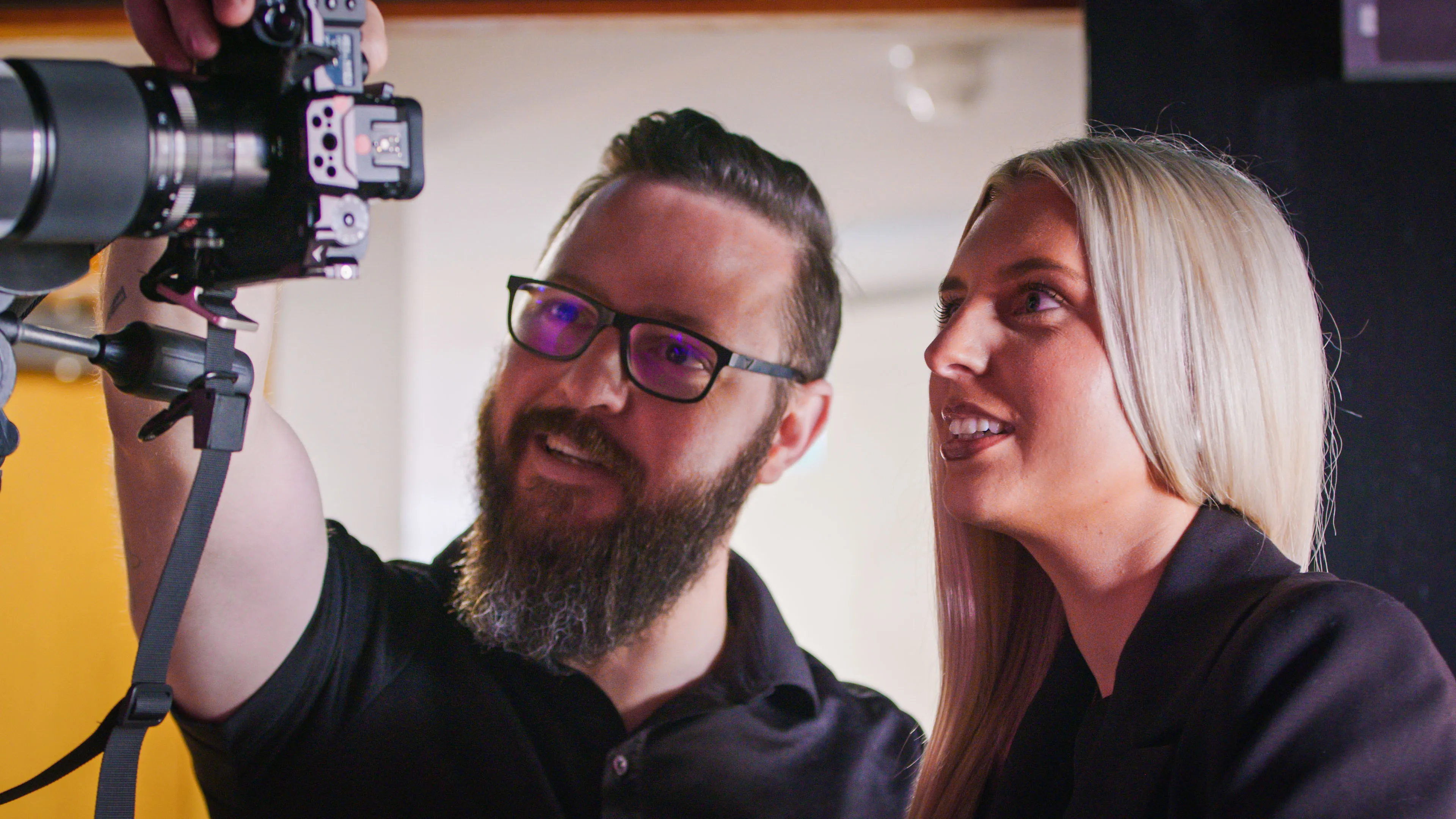 A bearded man wearing glasses and a smiling woman are looking at a camera on a tripod. The scene conveys a collaborative and focused atmosphere.