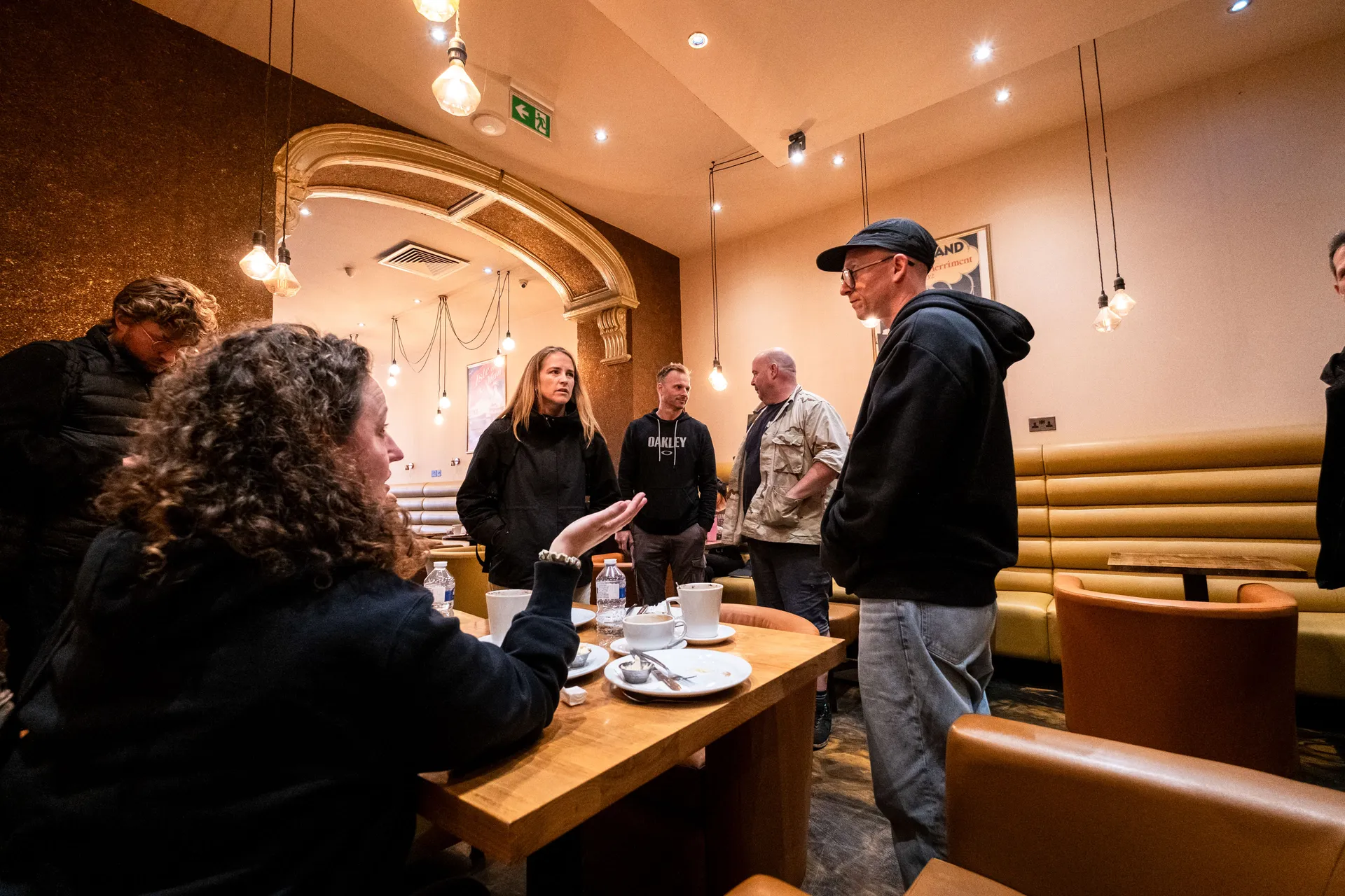 A group of people are gathered in a cozy cafe with warm lighting and arch decor. They seem engaged in a conversation, creating a friendly atmosphere.