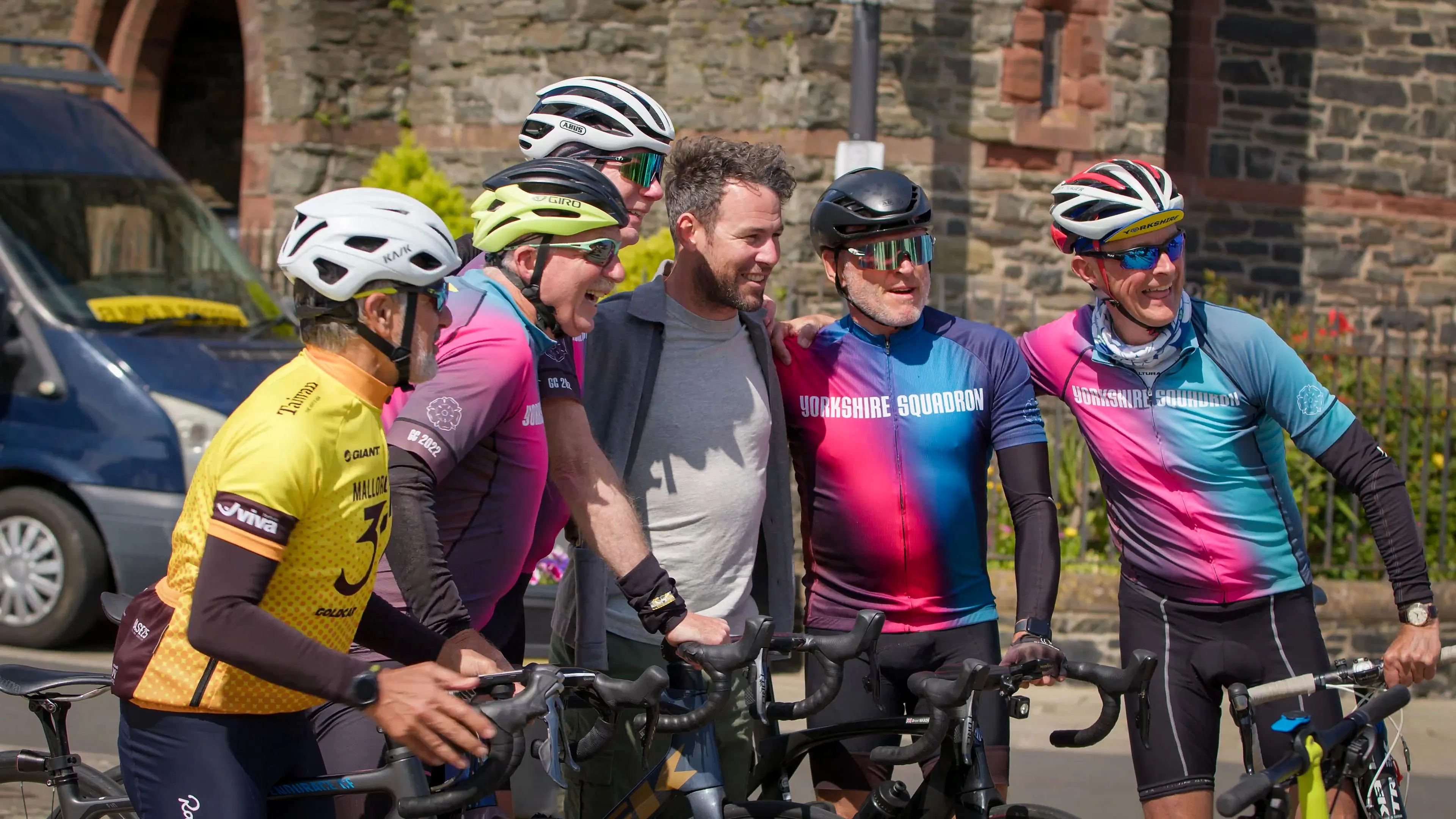 A smiling group of cyclists wearing colorful jerseys and helmets gather around a man in casual clothes smiling at a camera. They hold bikes on a sunny day by a stone building.