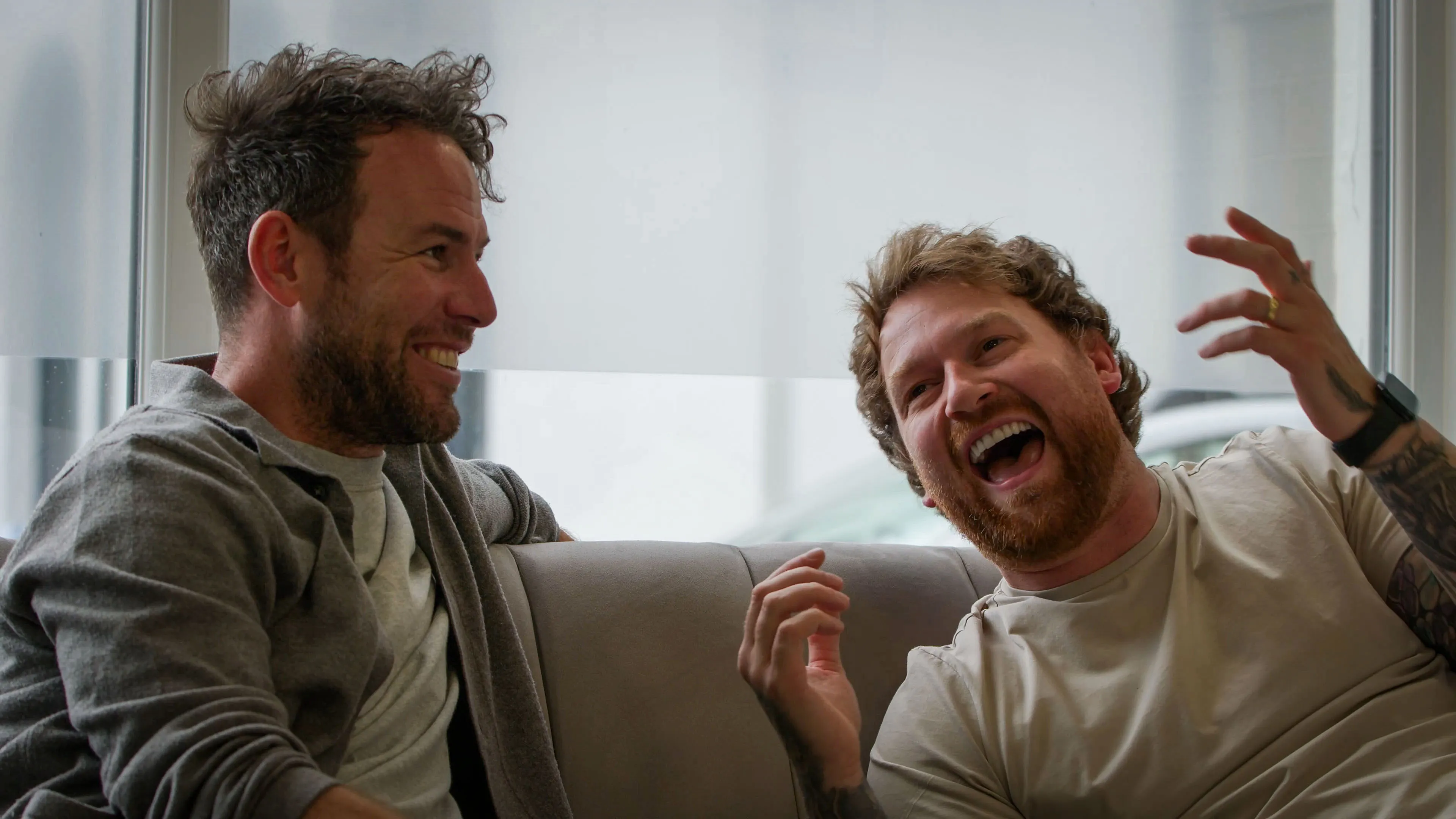 Two men sit on a sofa, laughing heartily. One wears a grey sweater, the other a beige shirt. The scene conveys joy and camaraderie in a bright room.