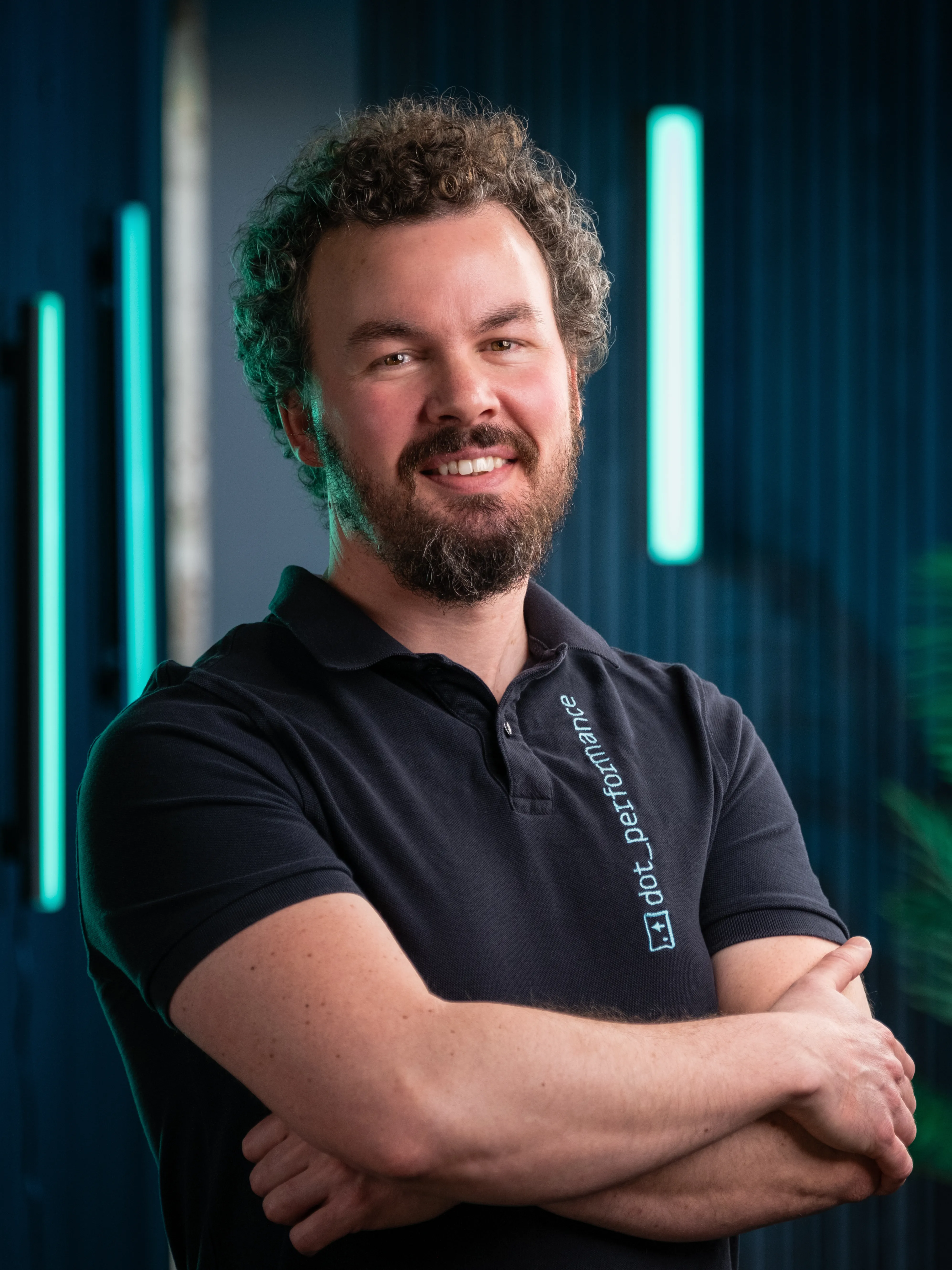 Adrian, UI/UX specialist at Dot Performance, smiling with crossed arms as he poses for headshot wearing branded hoody, against dark background with blue neon lights.