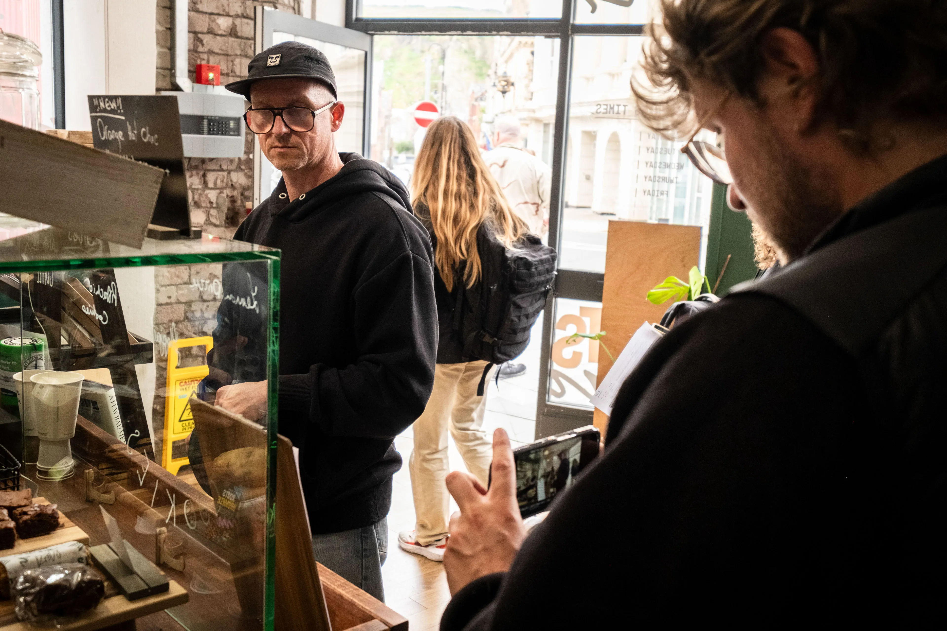 A man in a black hoodie and cap stands near a café counter with pastries. Another person films him on a phone. The atmosphere is casual and lively.
