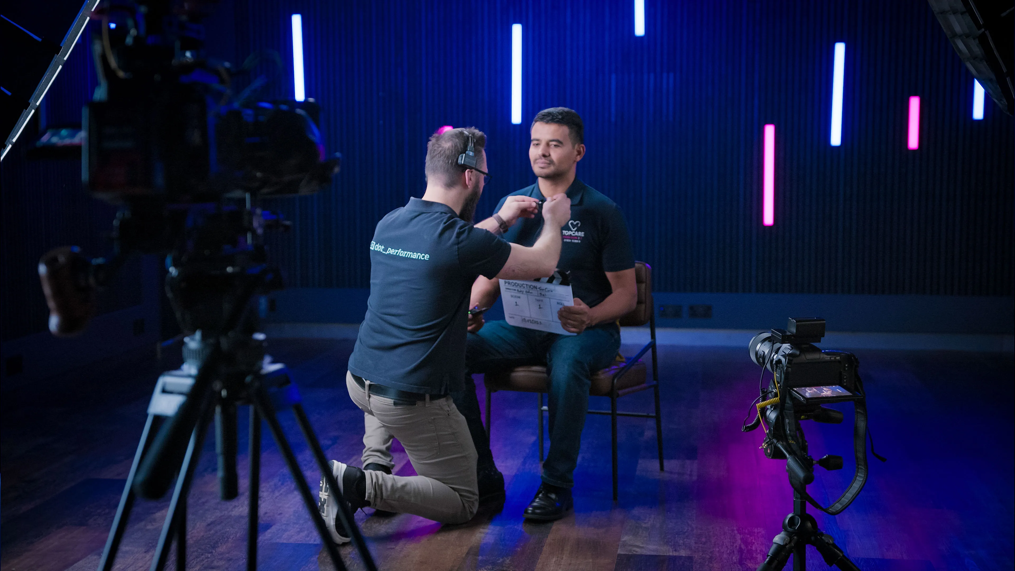 A technician adjusts a lapel microphone on a seated man in a dimly lit studio, with neon lights and cameras around, creating a professional atmosphere.