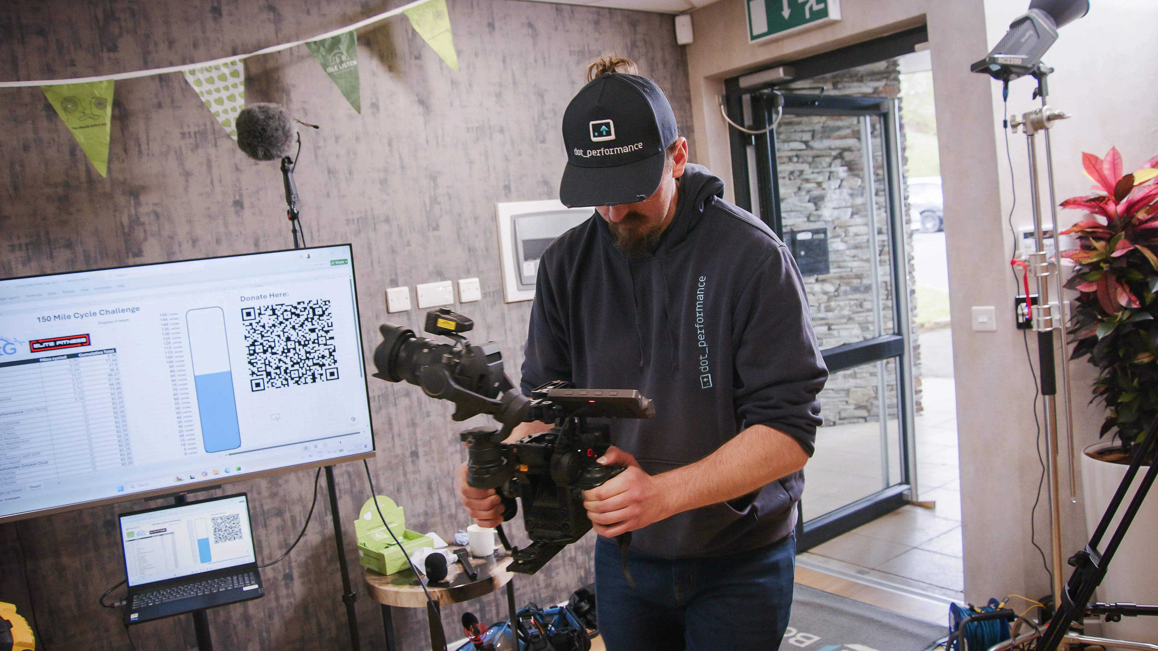 Will Oates from DotPerformance checking a DJI Ronin 4D cinema camera rig at the Boal & Co office on the Isle of Man, with a live fundraising tracker visible on a screen in the background