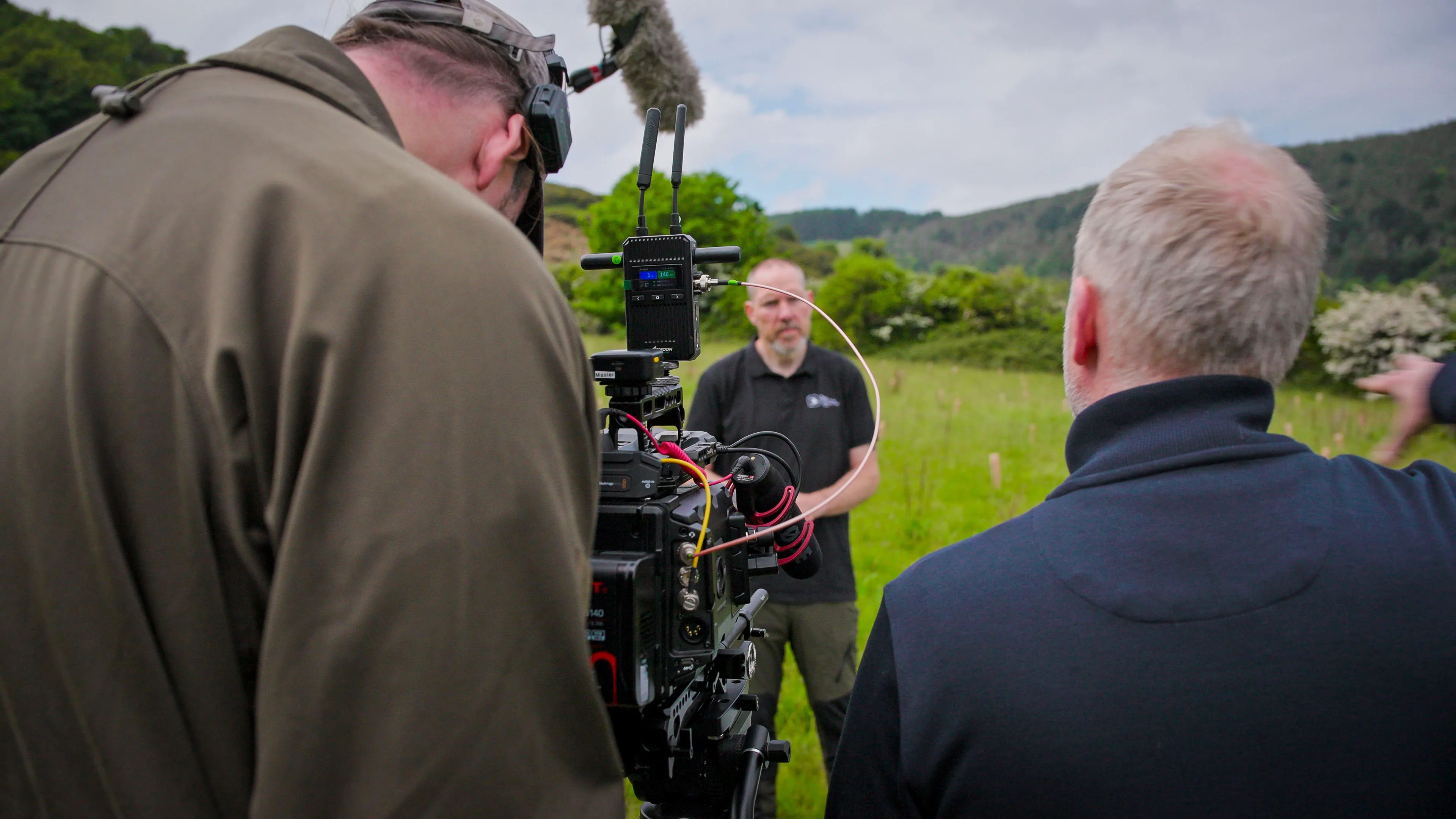 A cameraman films a man in a field with rolling hills in the background. The setup includes a large camera and microphone, creating a professional atmosphere.