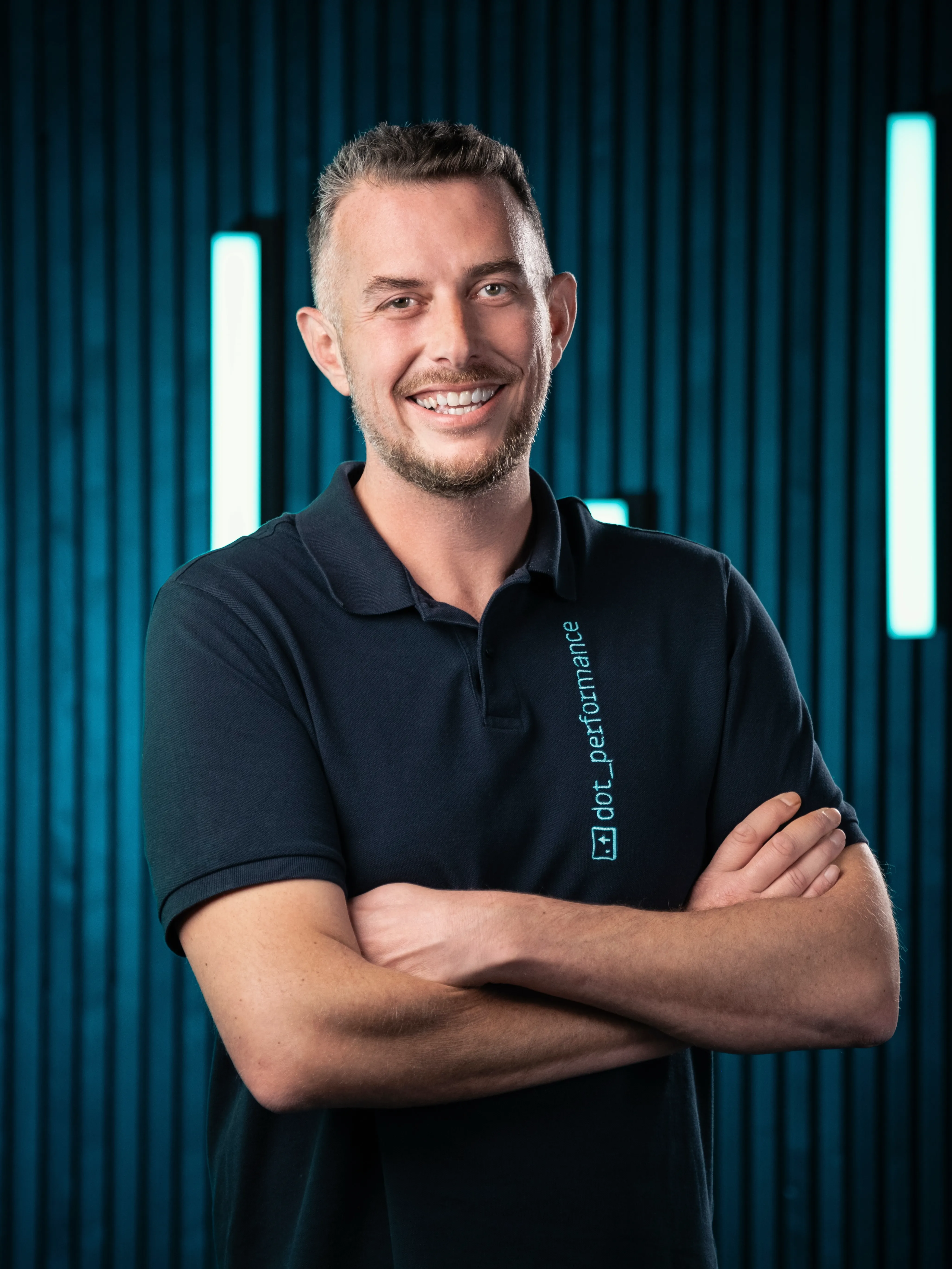 Andy Orton founder of DotPerformance, smiling wearing a DotPerformance branded polo shirt at DotPerformance studios on the Isle of Man, against a dark background with teal neon lighting