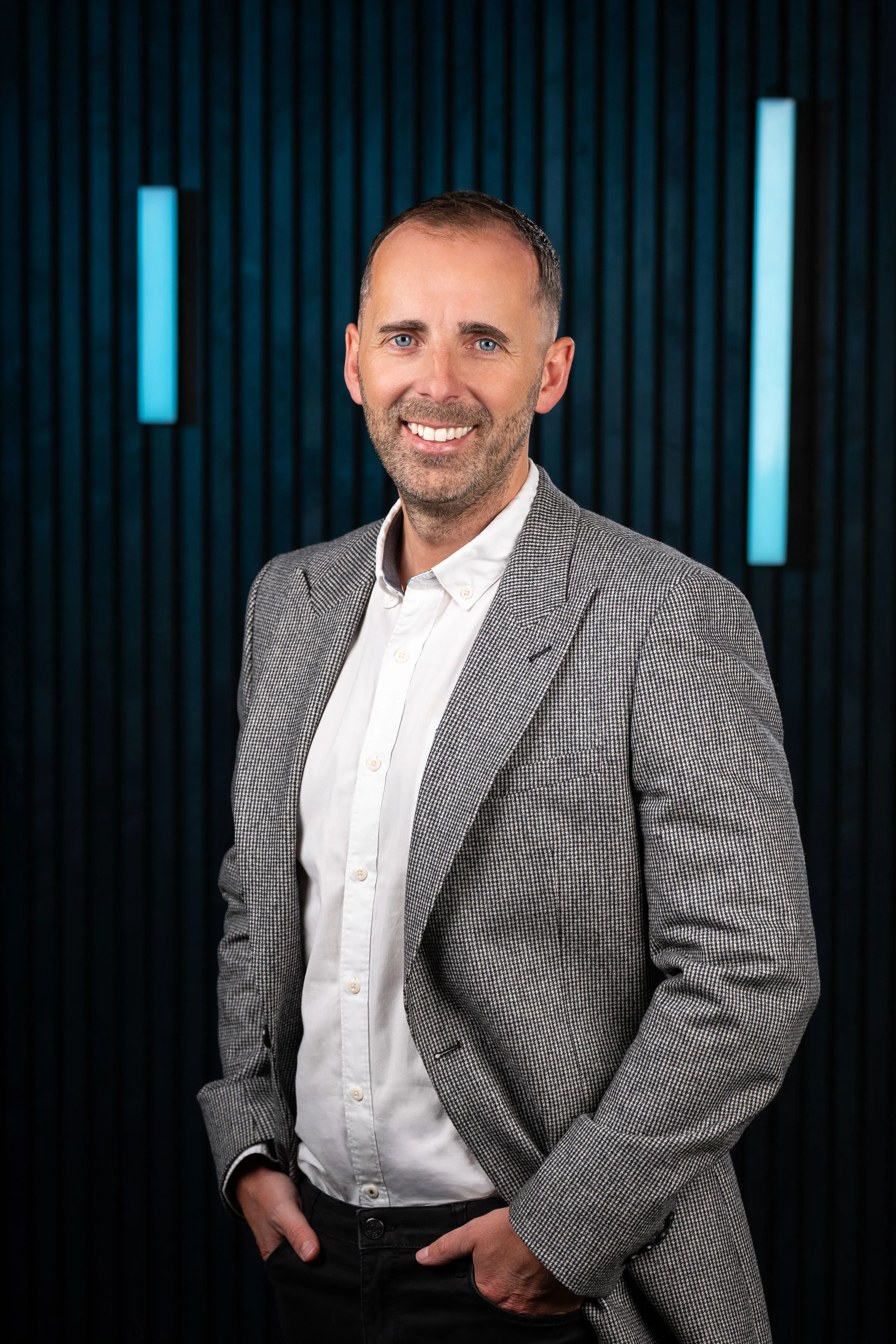 Man in a gray suit and white shirt, smiling with hands in pockets, stands against a dark, textured background with vertical blue lights. Professional and approachable tone.