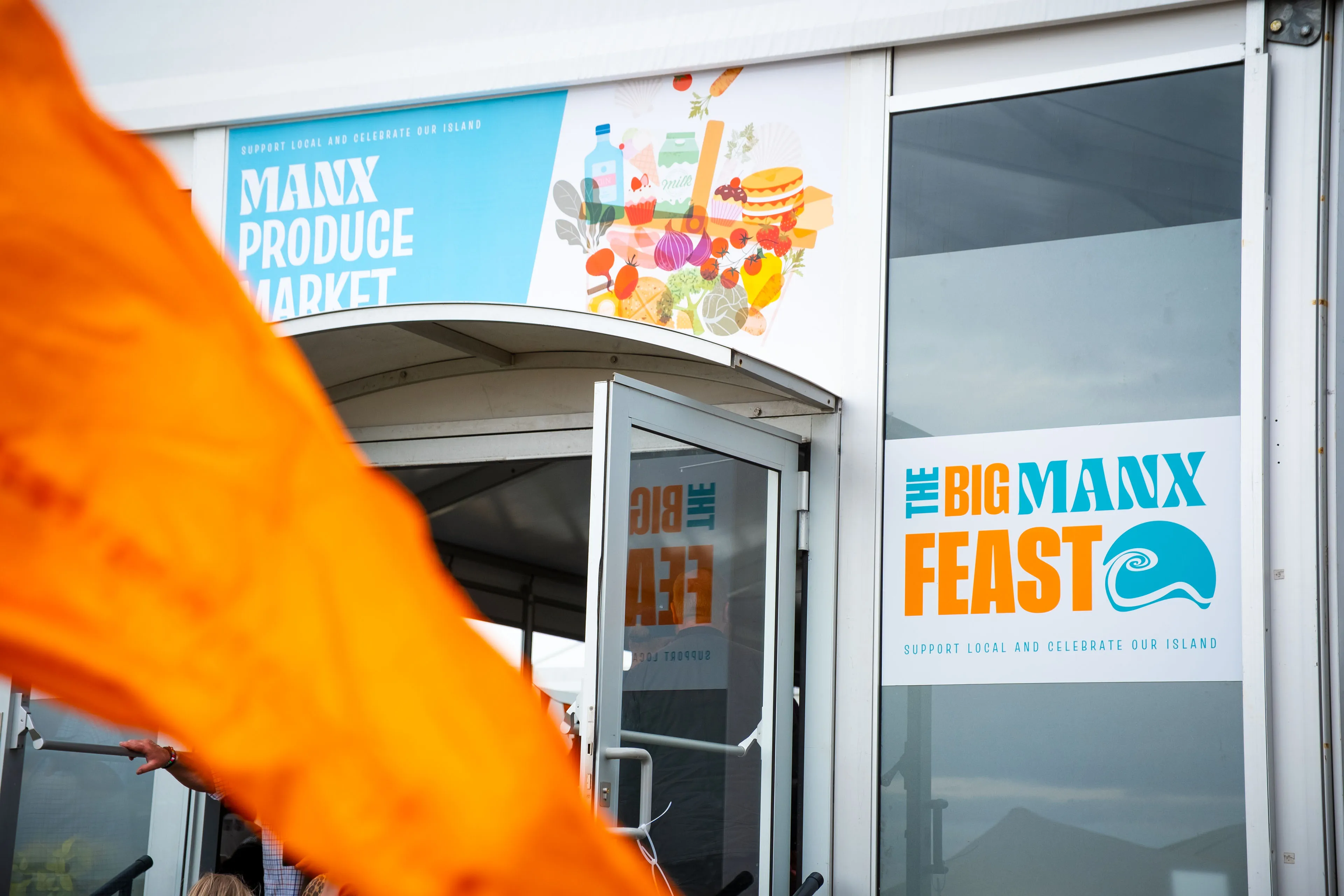 The Manx Produce Market hall entrance with Big Manx Feast branding, seen past an orange festival flag