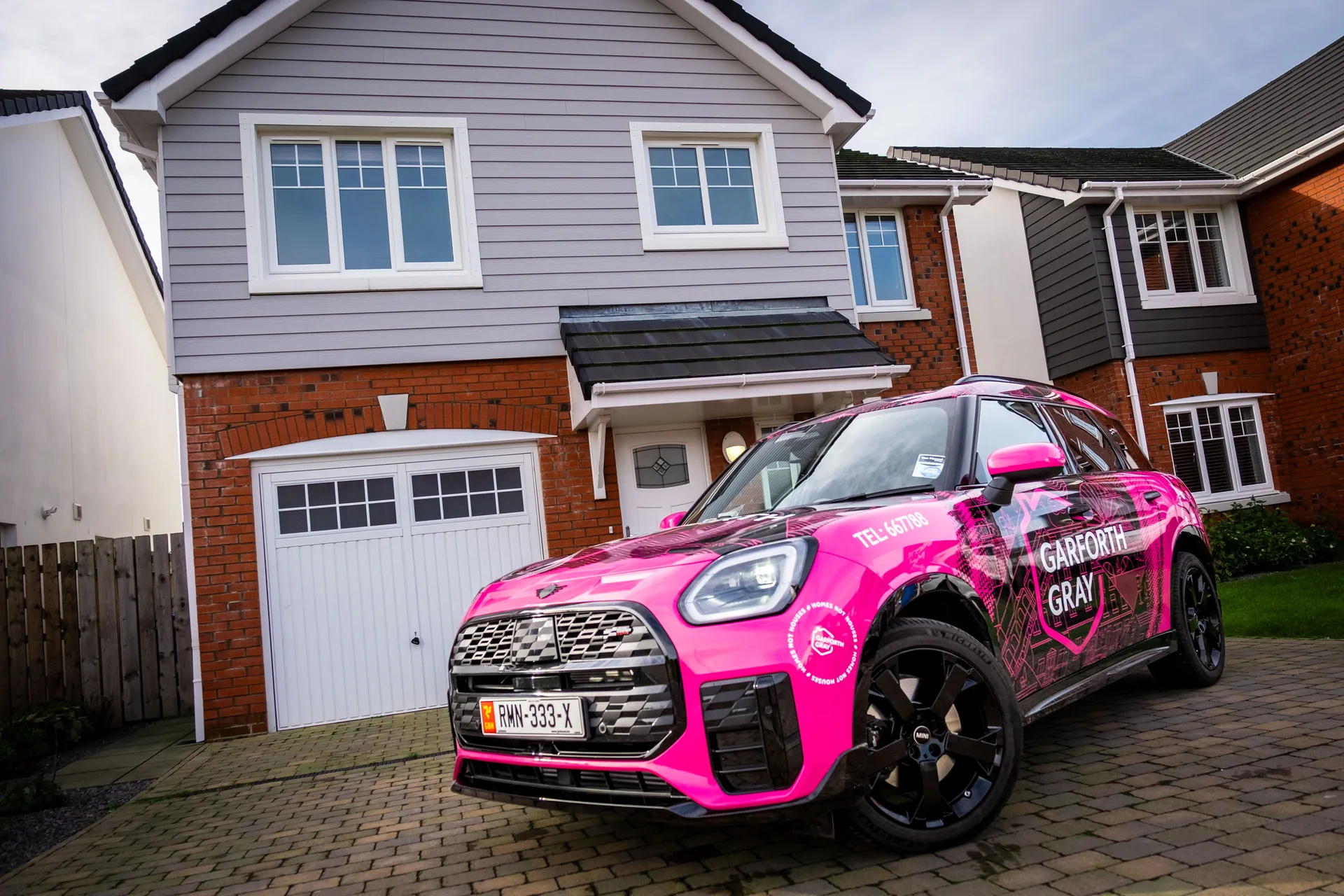 A bright pink Mini with black accents and "Garforth Gray" branding is parked on a brick driveway in front of a suburban two-story house.