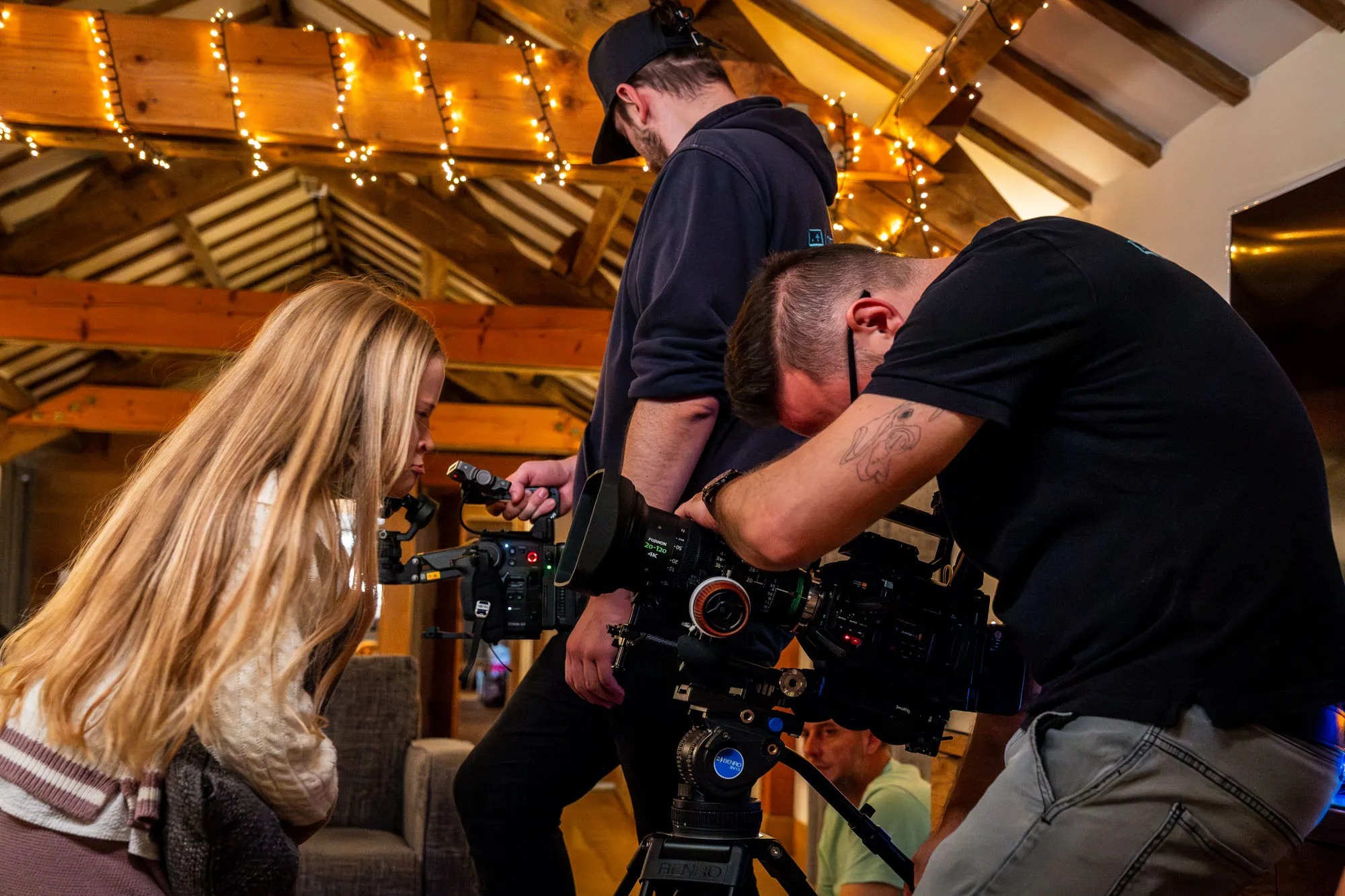 A cozy filming scene in a warmly lit wooden cabin. Two men with cameras and a girl with long blonde hair pulling faces into the camera. Creating a collaborative, creative atmosphere.