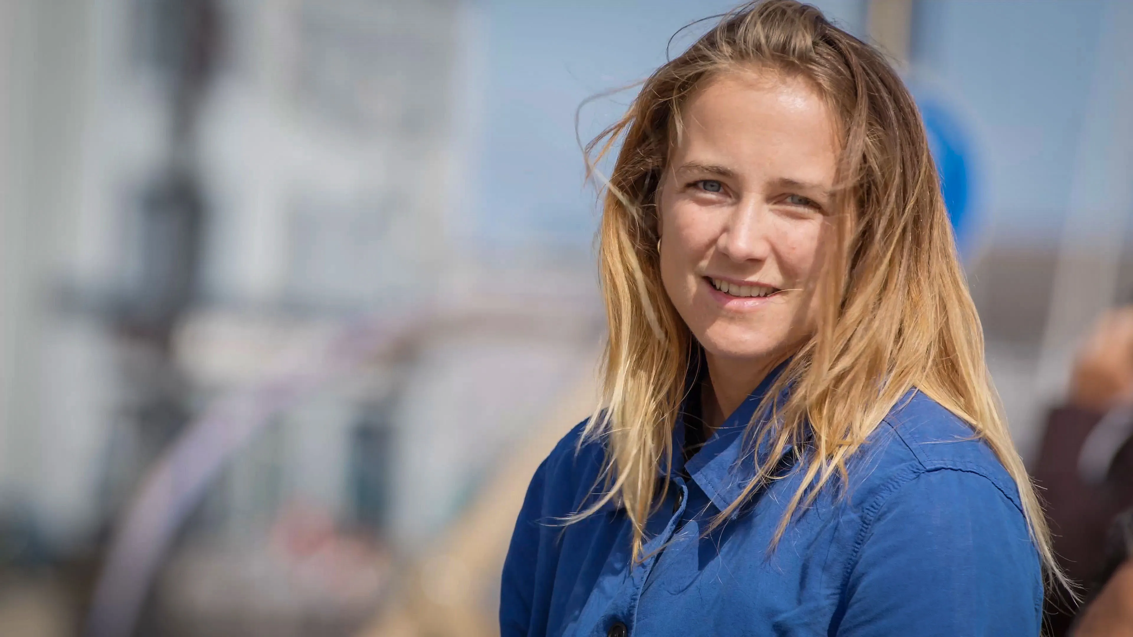Smiling woman in a blue shirt with windswept hair stands outdoors. The background is a blurred urban setting, conveying a casual and relaxed vibe.