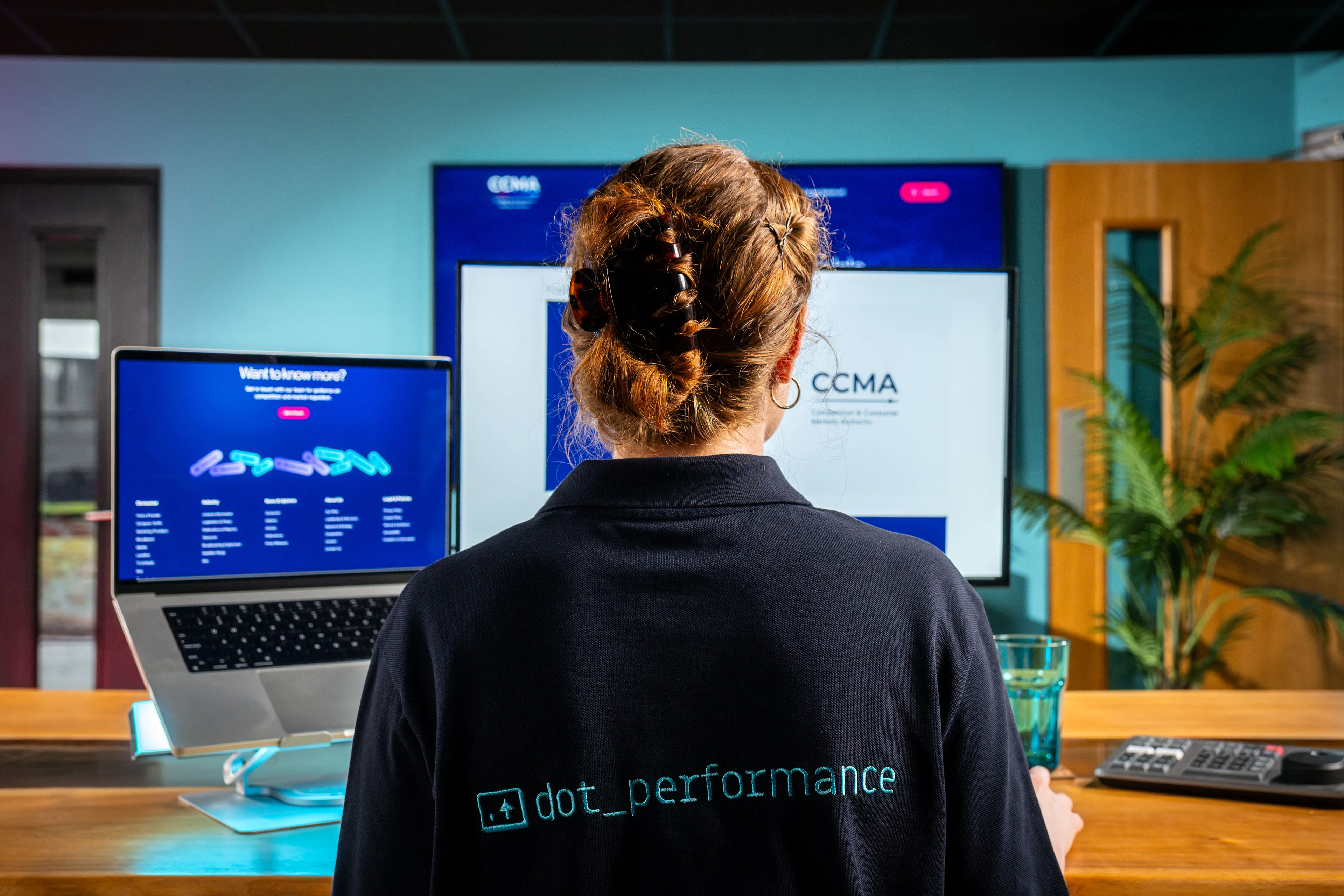 Over-the-shoulder view of Ella Garvey wearing a DotPerformance polo shirt while working on the CCMA website at DotPerformance