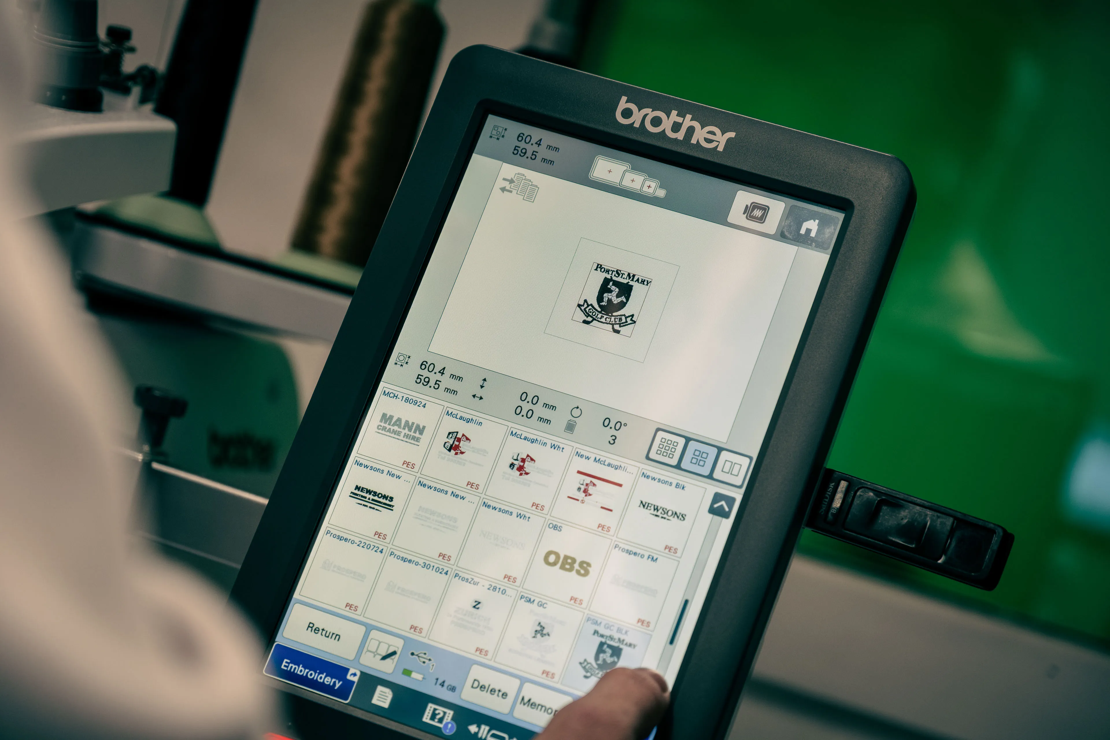 Close-up of a Brother embroidery machine screen showing a design preview. A hand operates the touchscreen, set against a vibrant green background.