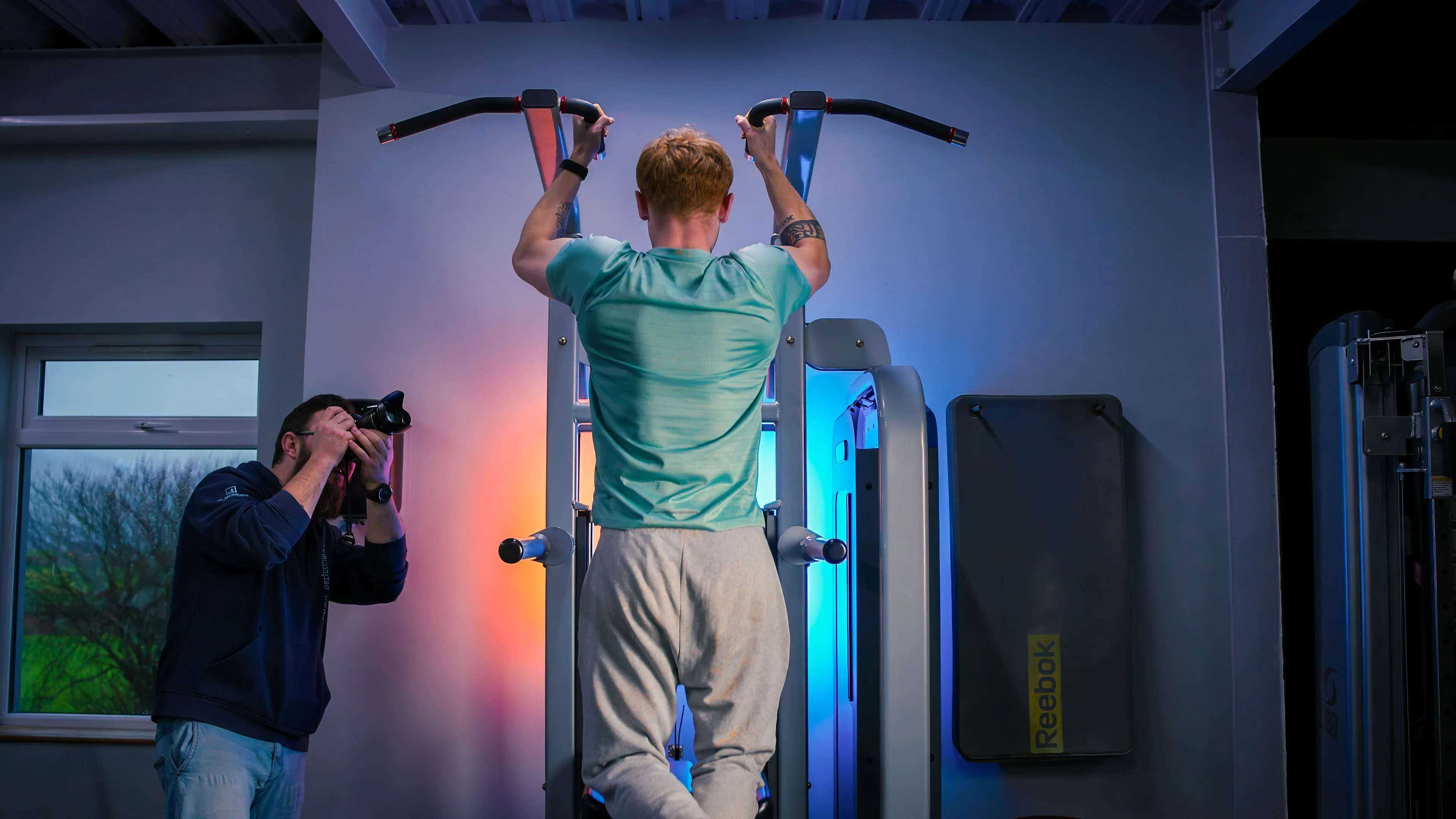 A person in a teal shirt performs a pull-up on gym equipment, while another person photographs them. The scene is lit with blue and orange hues.
