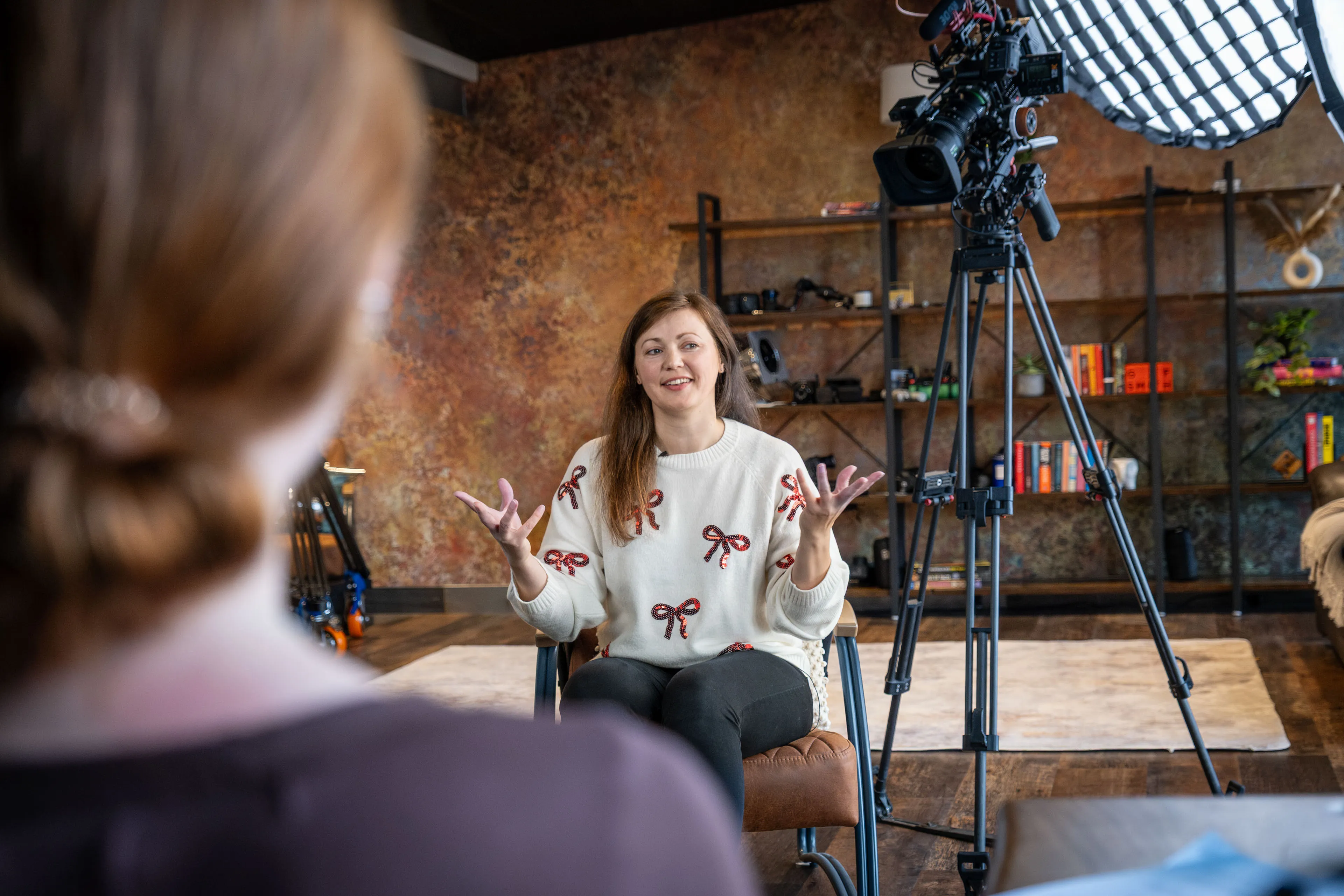 A person speaks animatedly in an interview setting, sitting in front of a camera with lighting equipment. A rustic wall and shelves form the background, creating a warm ambiance.