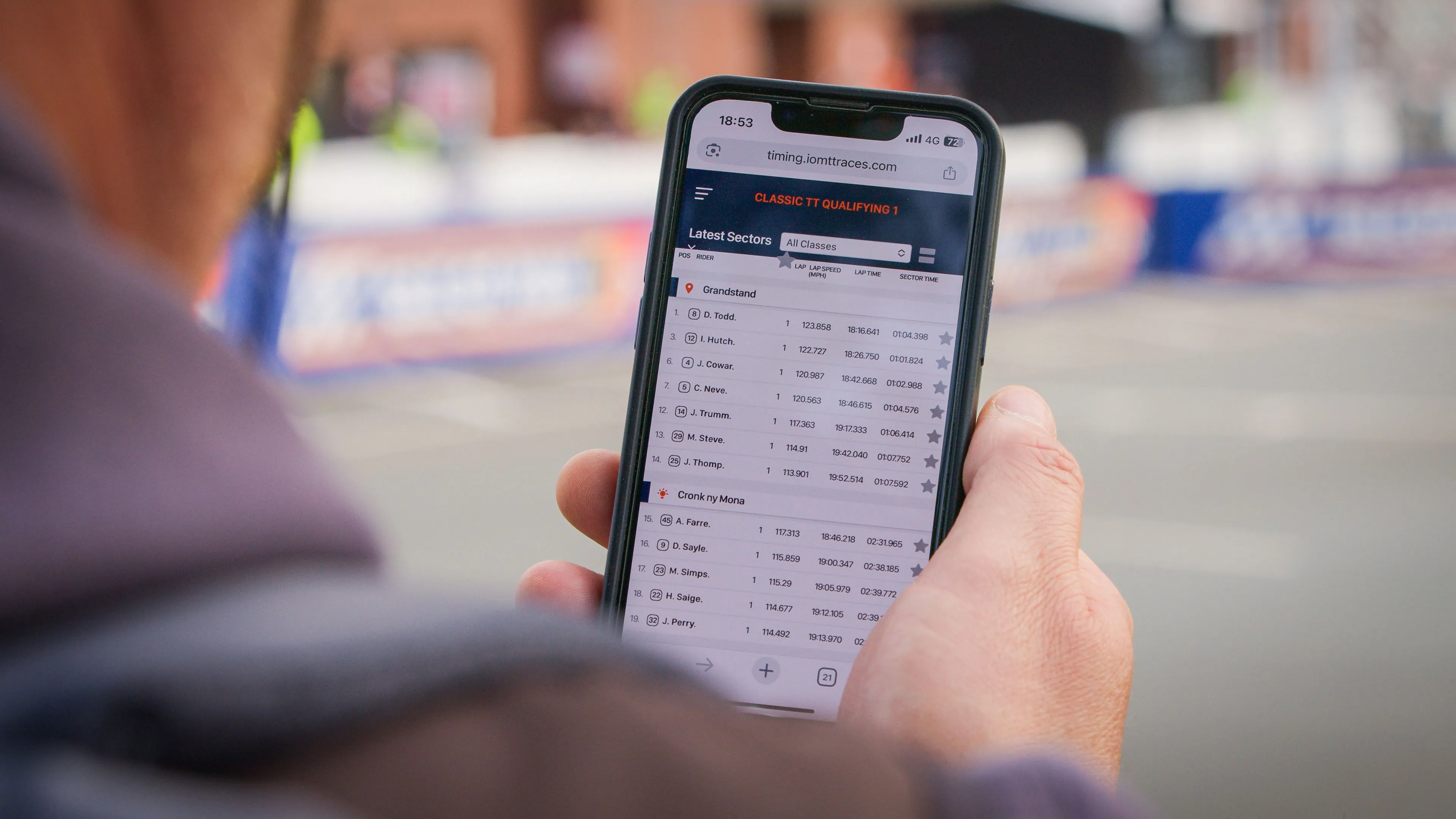 A spectator holding a smartphone displaying the DotPerformance Isle of Man TT live timing platform at timing.iomttraces.com, showing Classic TT Qualifying 1 sector times trackside at the Isle of Man