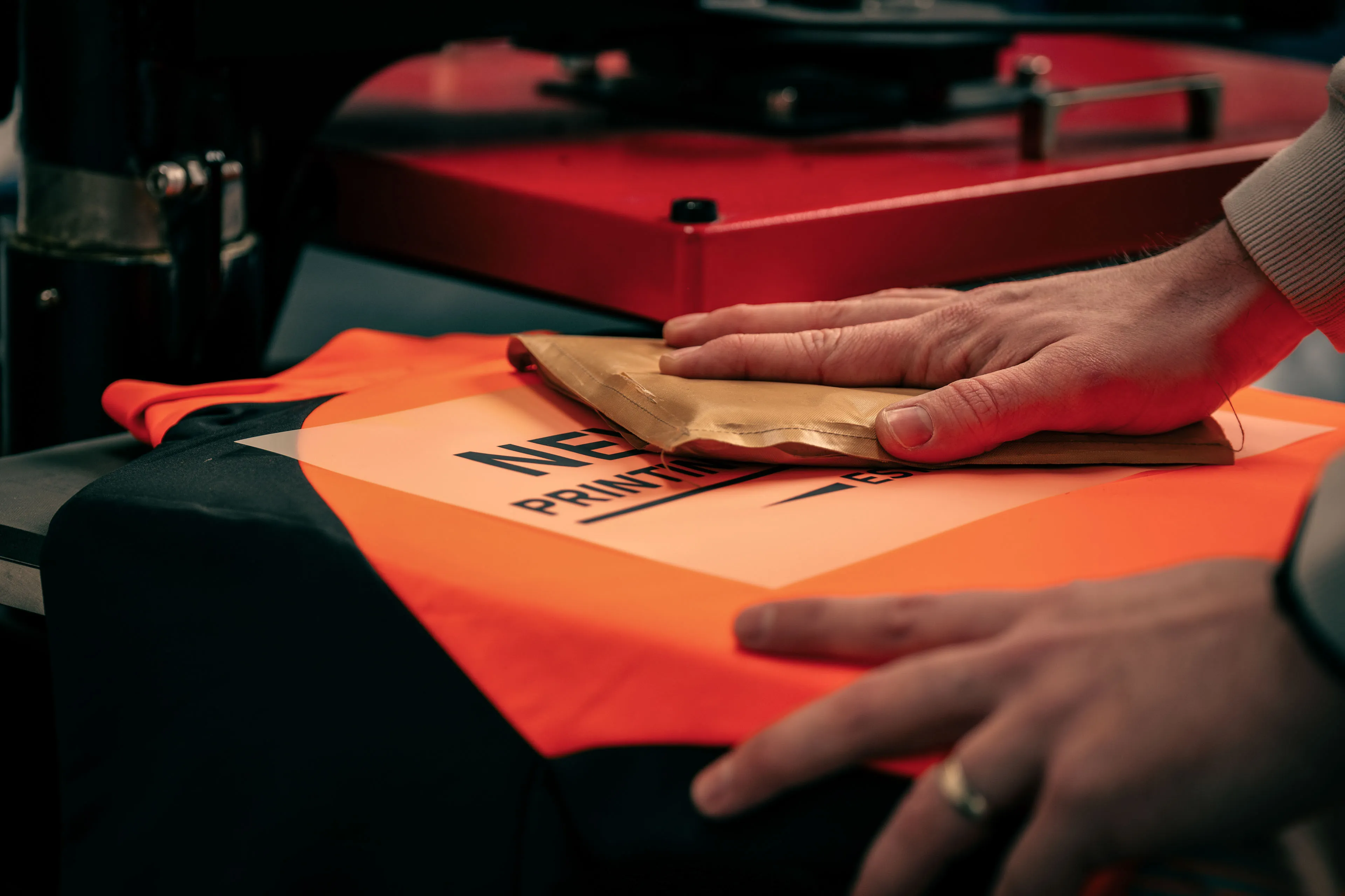 Close-up of hands smoothing a vibrant orange jersey with a brown paper on a heat press machine, focusing on printing details. The scene conveys precision and craftsmanship.