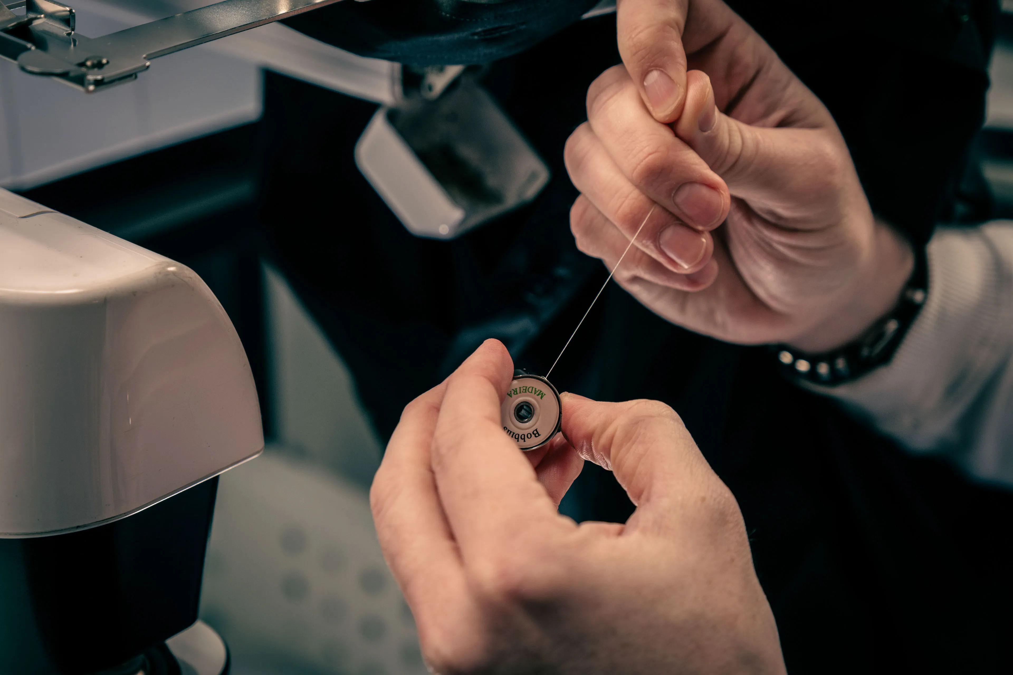 Close-up of hands loading black thread onto a bobbin, with sewing machine parts visible in the background. The scene conveys focus and precision.