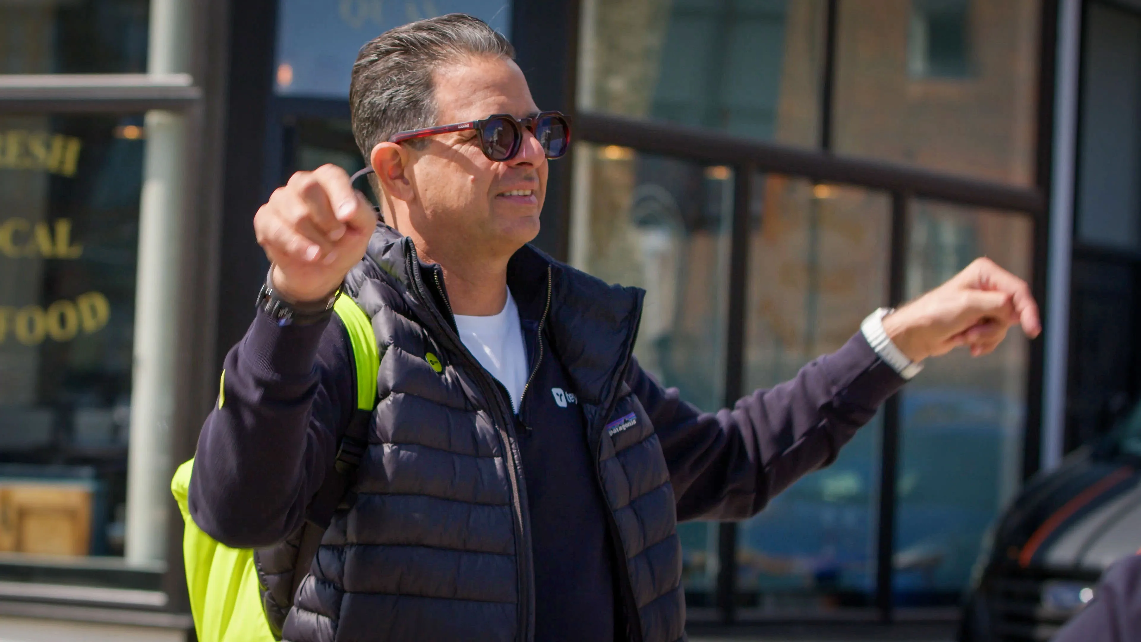 A man in sunglasses smiles and gestures with his arms raised outdoors. He wears a puffy jacket and has a neon green backpack. The mood is cheerful and lively.