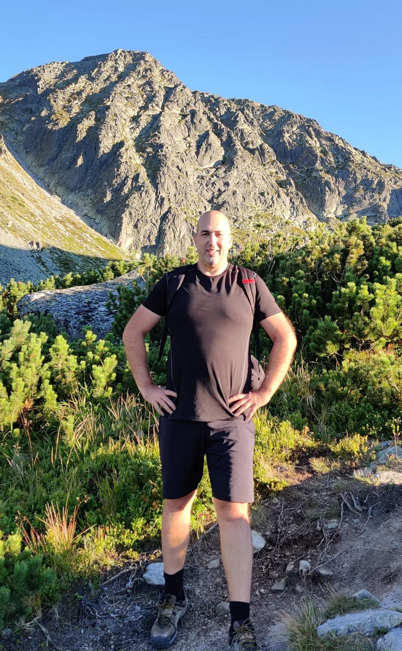 Claudiu Danila standing with hands on hips smiling on a mountain trail, with rocky peaks andEastern European vegetation in the background
