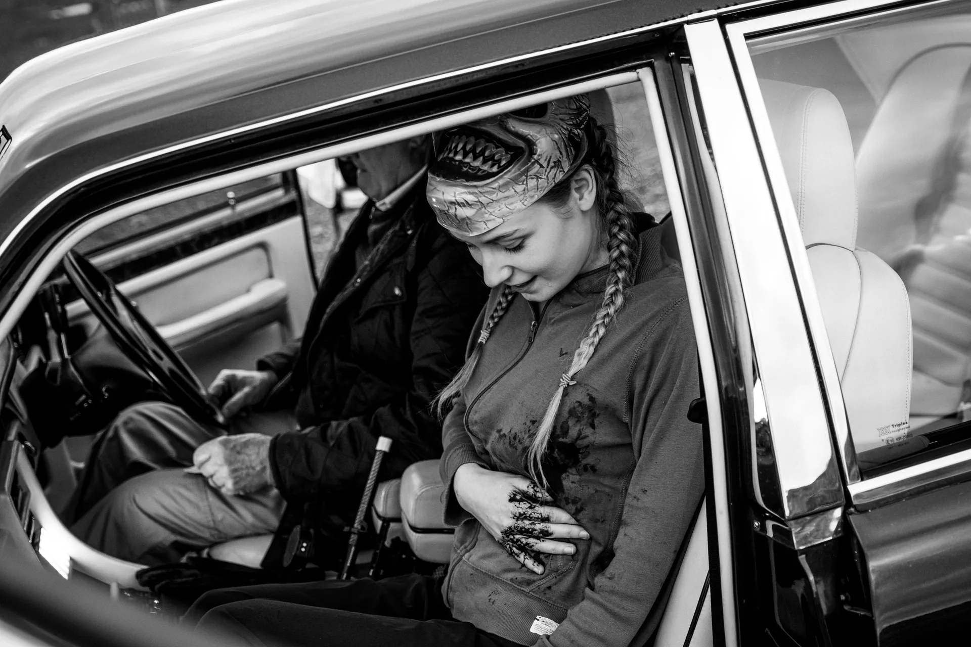 A young woman with braided hair and a mask on her head sits in a car's passenger seat, looking down at her bloody hand and abdomen with concern. A man sits in the driver's seat. The scene conveys tension.