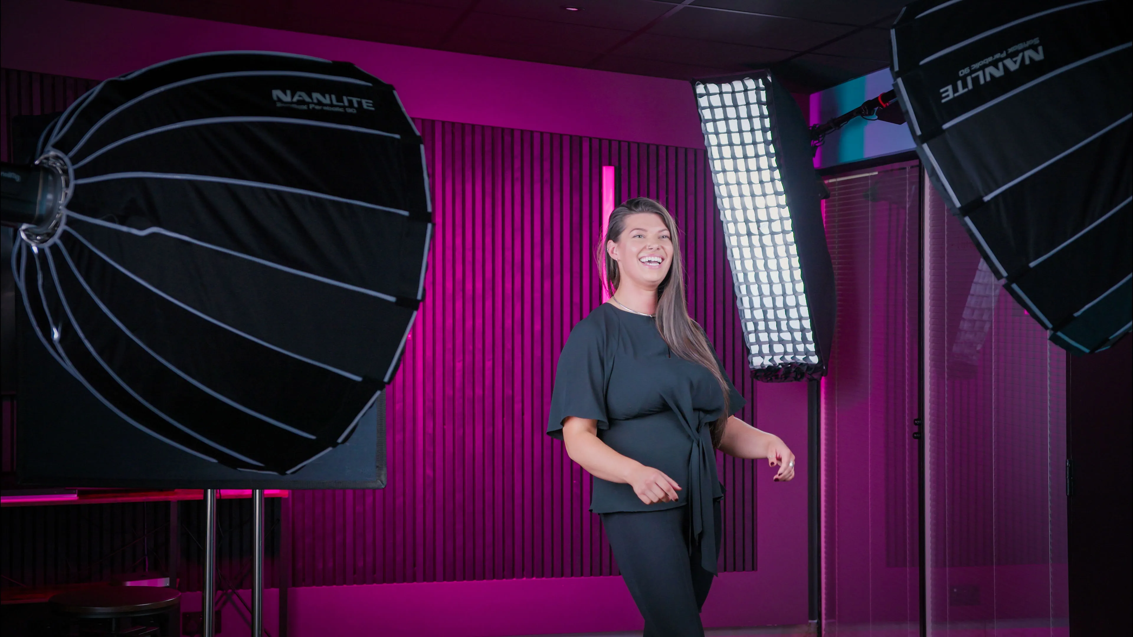 A woman in a studio with professional lighting smiles warmly. The background features vibrant pink walls and vertical lines, creating a lively atmosphere.