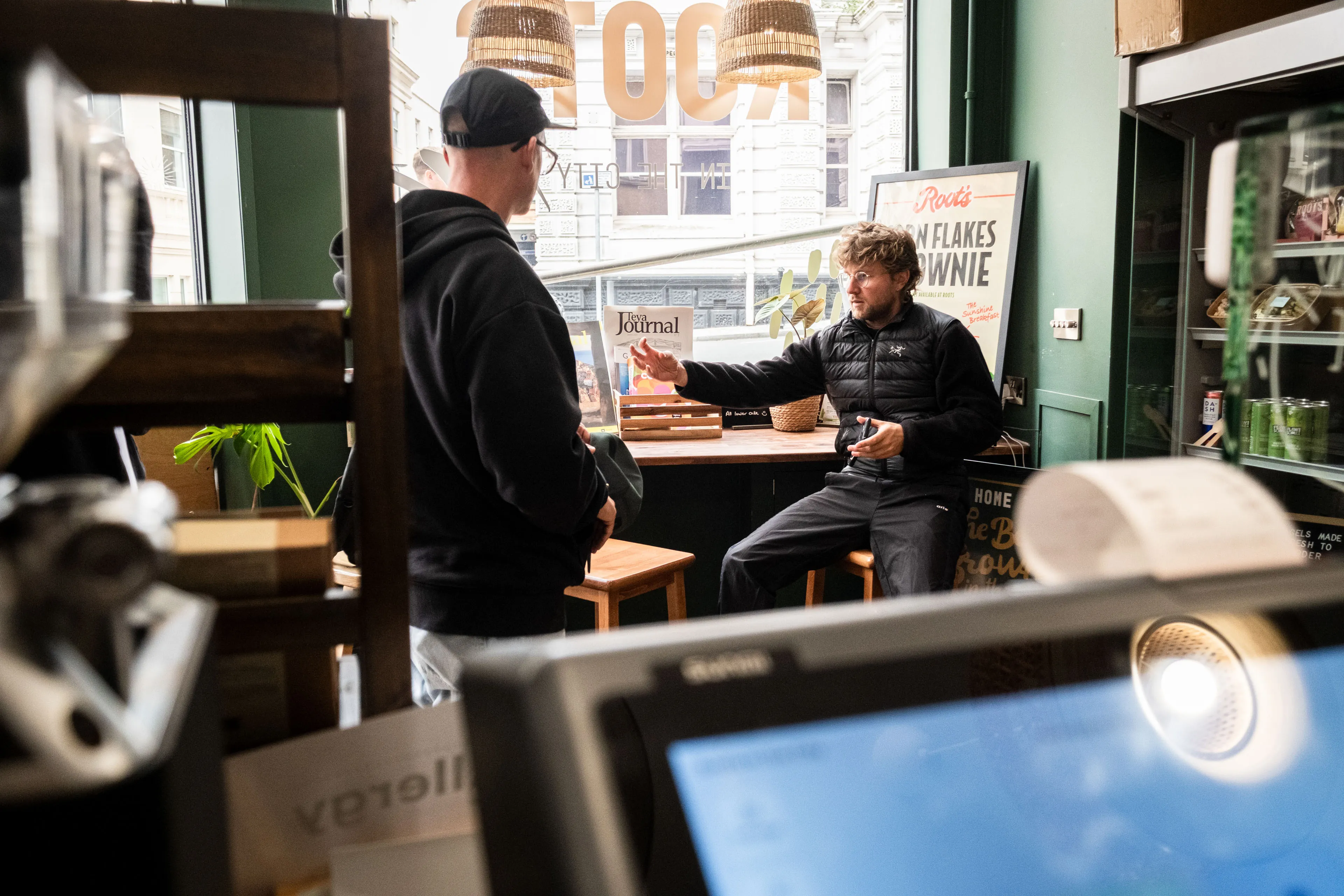 A man in a black jacket sits on a stool in a cozy café, gesturing while speaking to another man standing nearby. The atmosphere is casual and engaged.