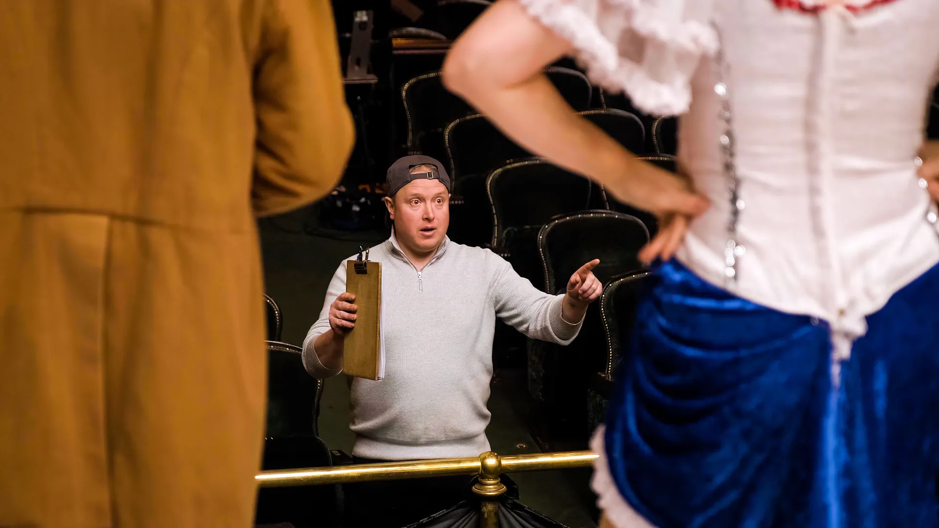 A director in a white sweater, holding a clipboard, gestures while instructing performers on stage. The scene conveys focus and teamwork.