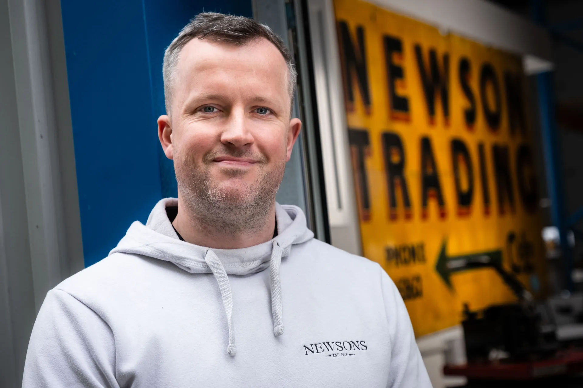 A man in a light beige hoodie stands confidently in front of an industrial building, with a blue wall and a yellow vintage "Newsons Trading" sign.