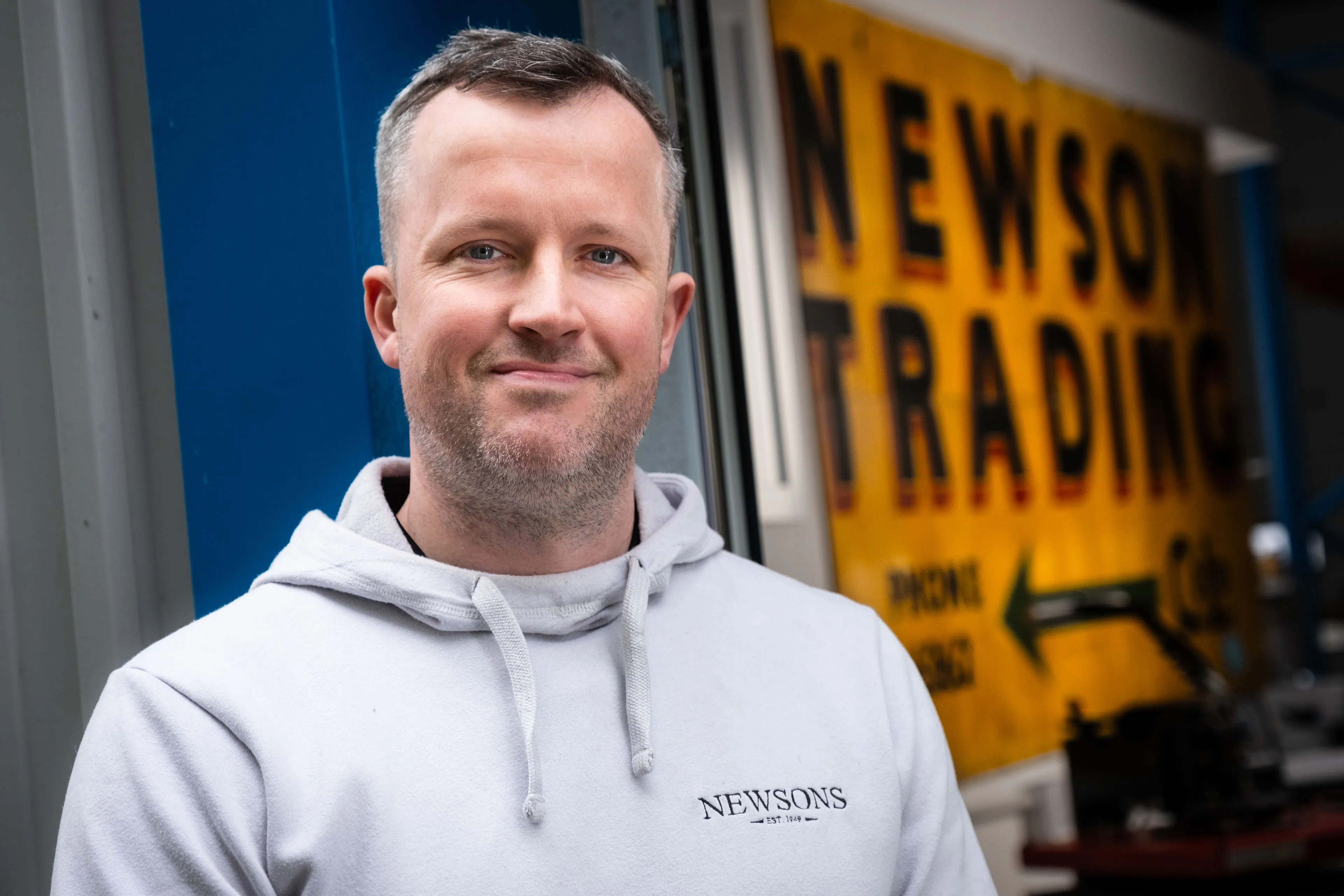 A man in a light beige hoodie stands confidently in front of an industrial building, with a blue wall and a yellow vintage "Newsons Trading" sign.