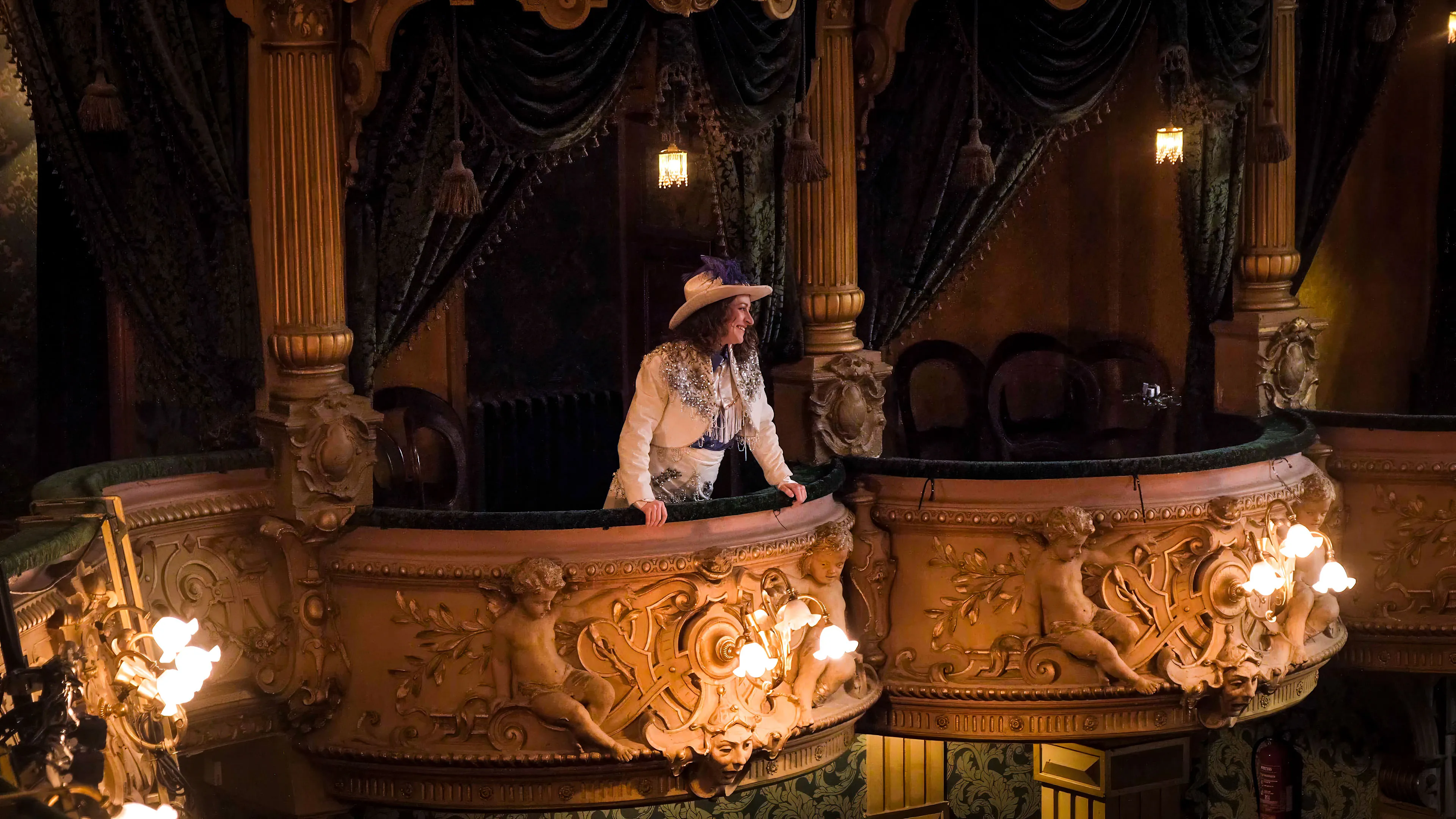 A person dressed in period costume stands on a lavish theater balcony, decorated with ornate carvings and draped dark green curtains, under warm lighting.