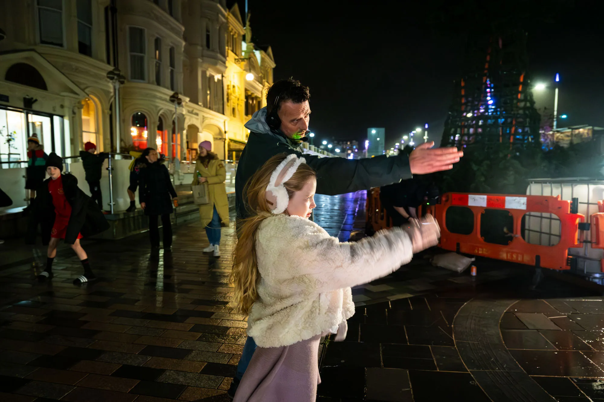 A man and a young girl in earmuffs gesture energetically on a lively nighttime street, surrounded by people and illuminated buildings, creating an animated and joyful atmosphere.