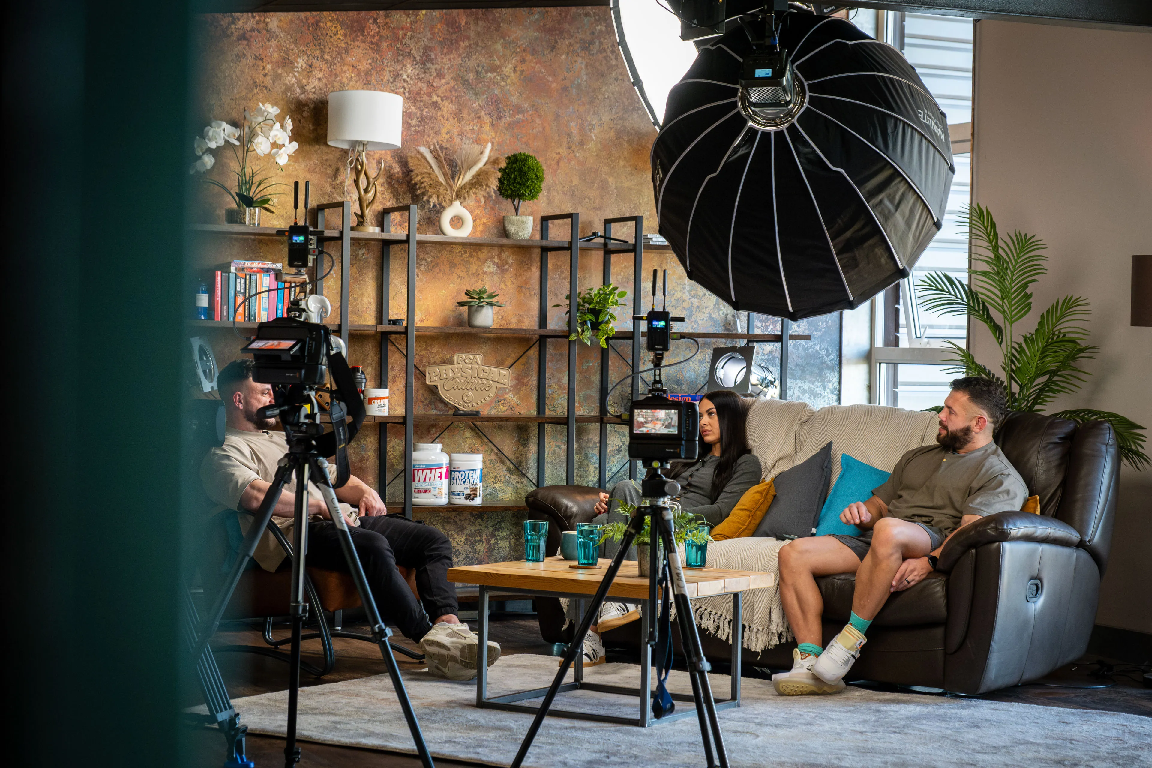 Danny and Christie Hands seated on a sofa talking to Jake Watts in the DotPerformance podcast studio on the Isle of Man, with cinema cameras and Nanlite lighting visible in the foreground during a multi-camera interview recording