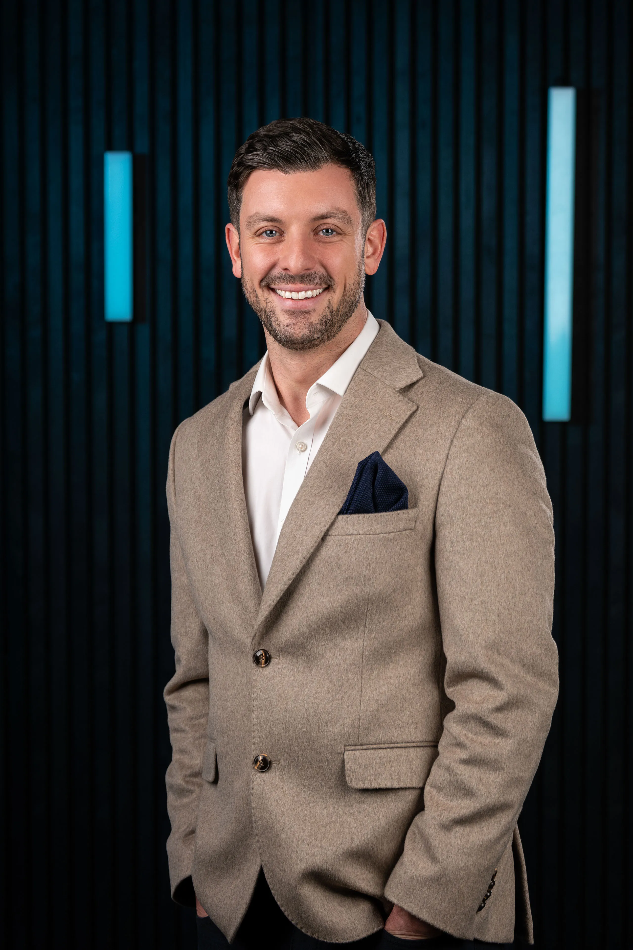 Professional headshot of Dan Collier smiling, wearing a tan blazer and white shirt with a navy pocket square, against a dark studio backdrop with blue accent lighting