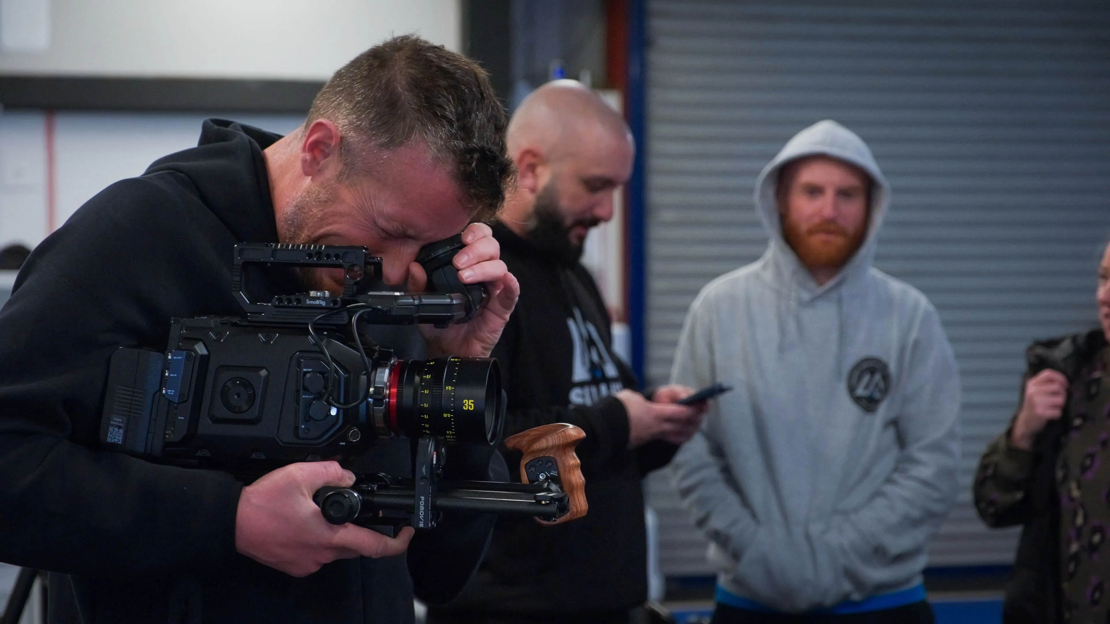 A man focuses intently through a professional camera, while two others stand in the background; one uses a phone, the other observes, creating a relaxed, collaborative atmosphere.