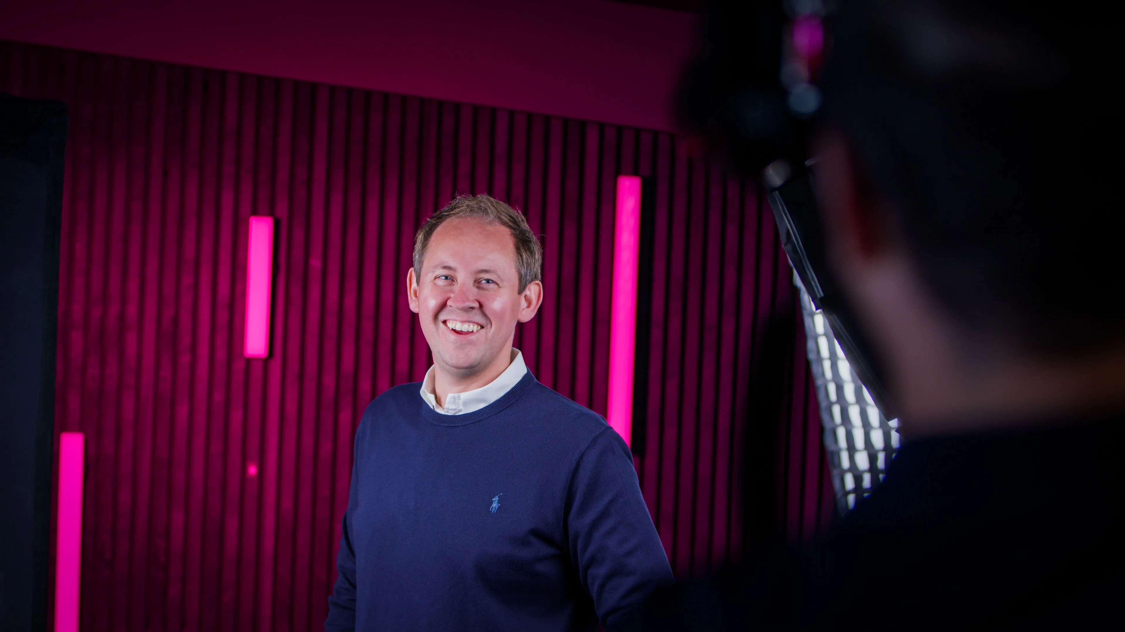 Man in a blue sweater smiles under bright studio lights, framed by a magenta textured wall with vertical light bars, creating a vibrant, upbeat atmosphere.