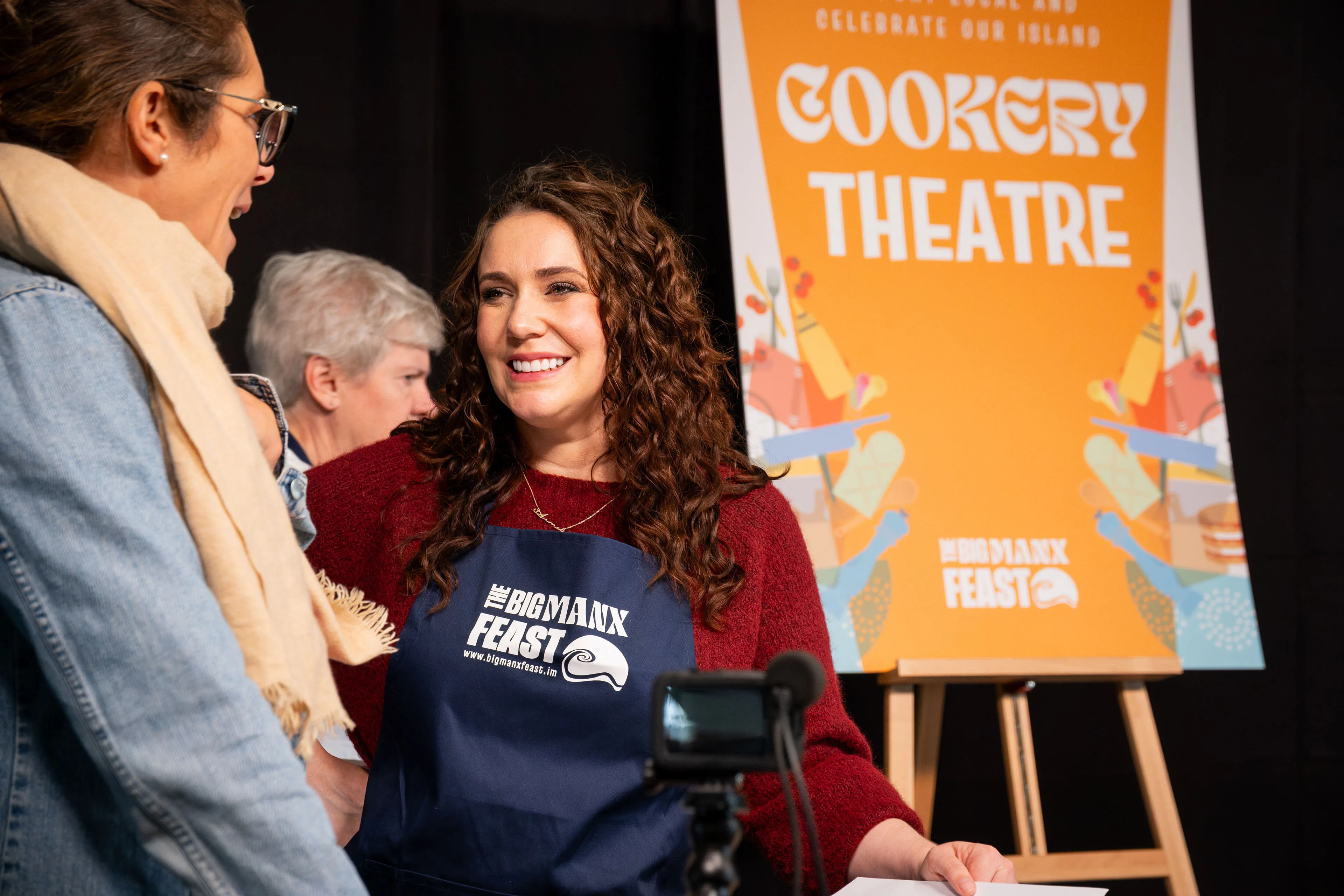 A chef in a Big Manx Feast apron talking with attendees in front of the Cookery Theatre sign