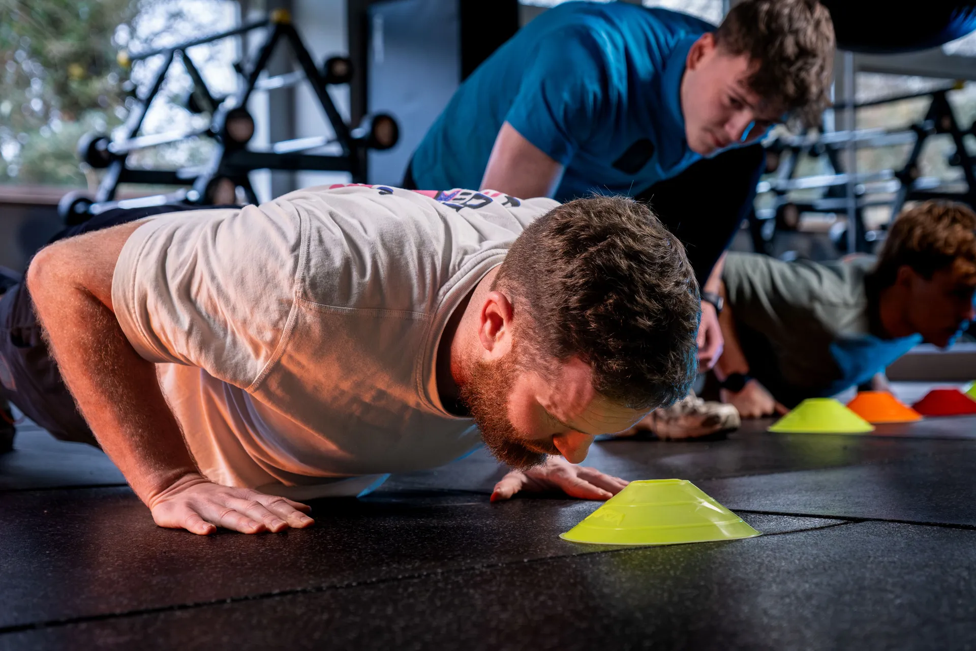 Three men performing push-ups in a gym, focused and determined. Coloured cones are placed on the floor as markers. Equipment is visible in the background.