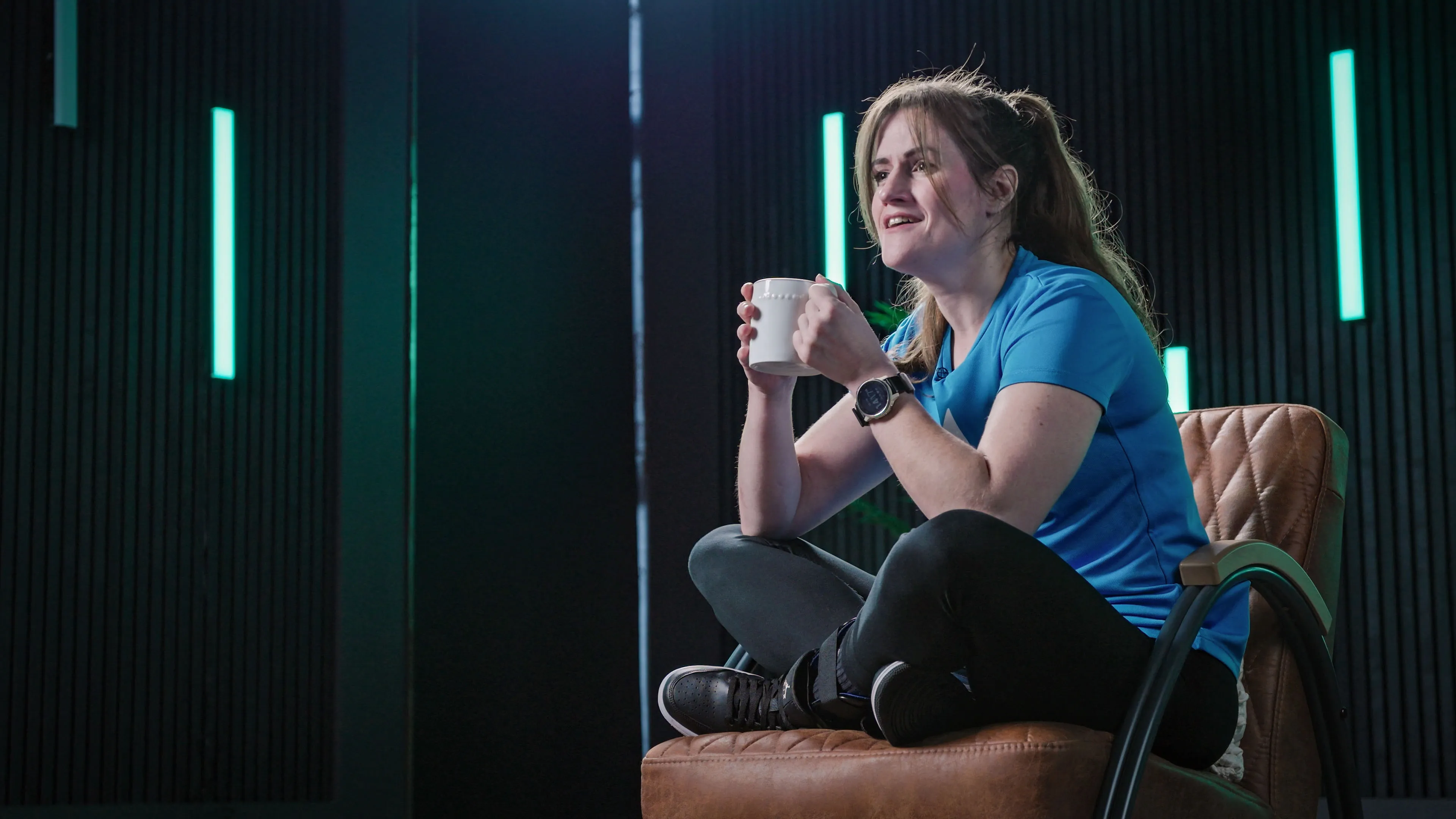 Woman in a blue shirt sits cross-legged on a brown chair, holding a mug. She smiles, exuding a relaxed mood. Neon lights in the background.
