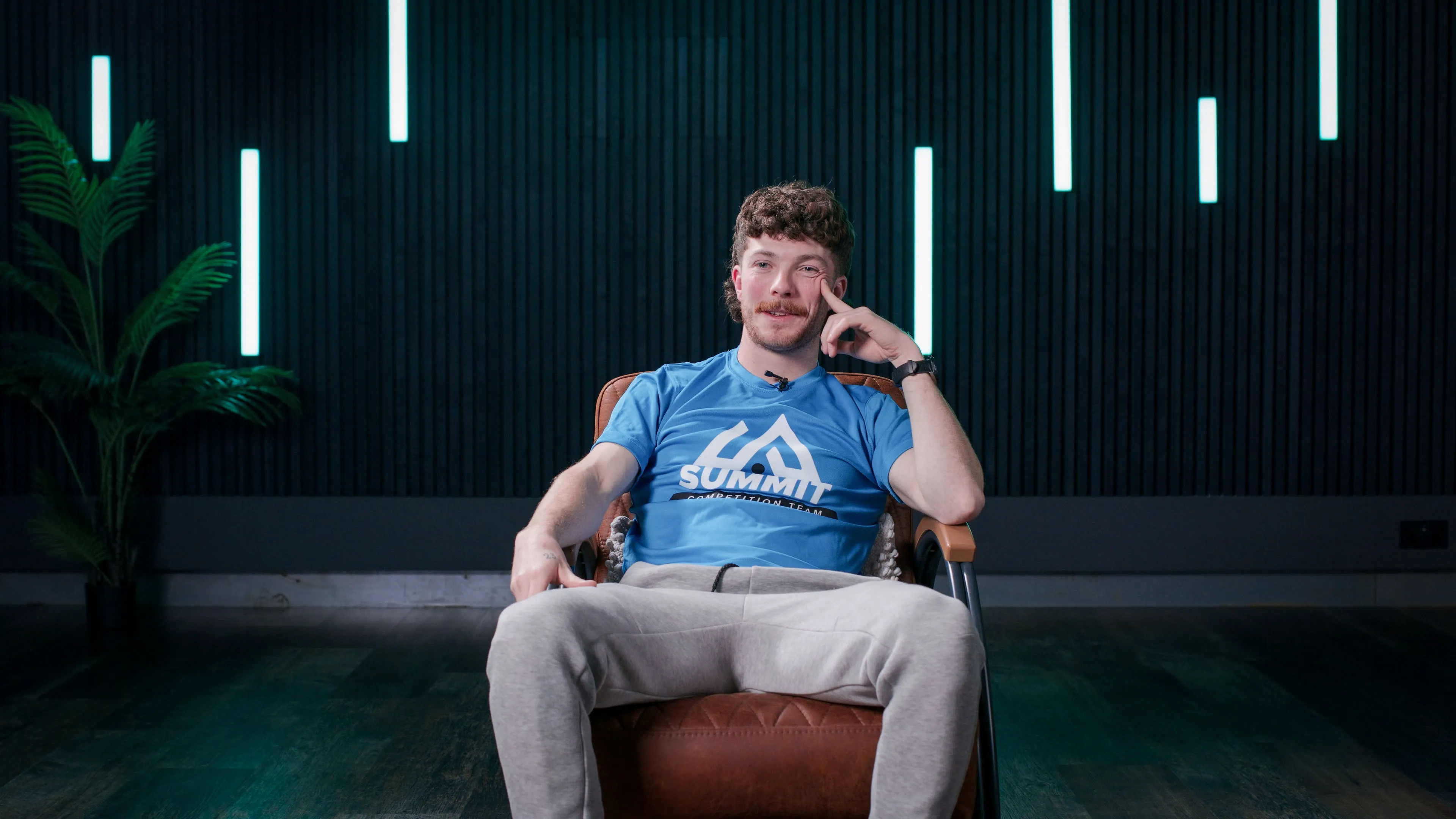 A person in a blue Summit T-shirt and gray pants sits relaxed in a brown chair. The background features vertical light strips and a potted plant, creating a modern and calm atmosphere.