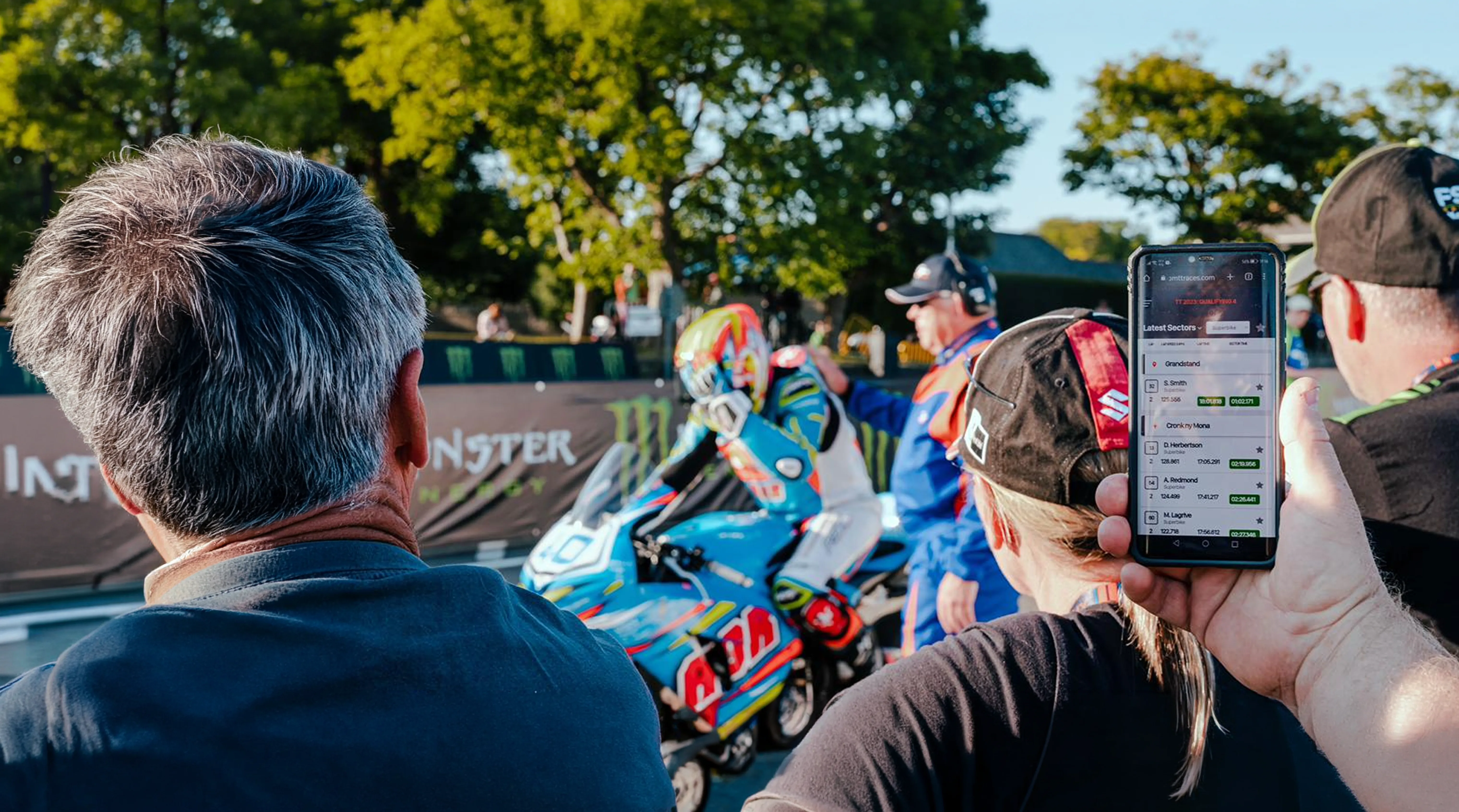 A spectator holds up a smartphone to film or photograph a motorcycle racer on a colourful race bike at the Isle of Man TT, with crowds and green trees visible in the background.
