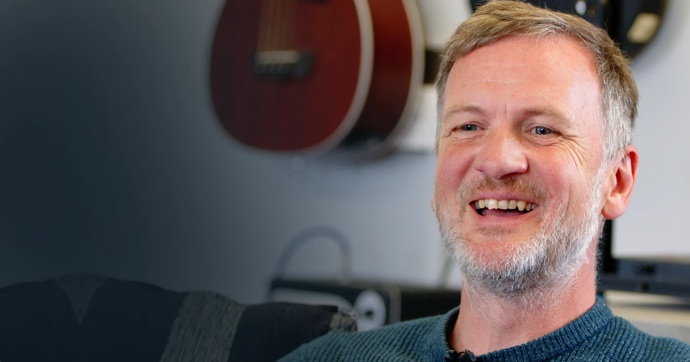 A smiling man with a gray beard and short hair is sitting in a room with guitars on the wall. He wears a blue sweater, conveying a relaxed and happy mood.