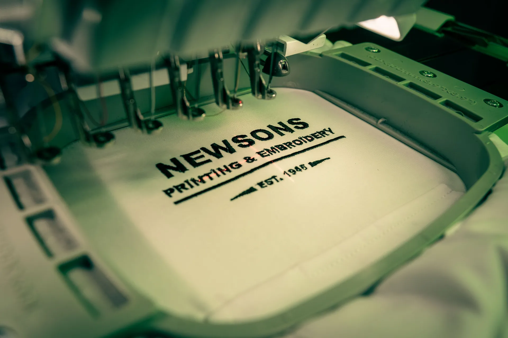 Close-up of an embroidery machine stitching "NEWSONS Printing & Embroidery, Est. 1985" on white fabric. The scene has a focused, professional tone.