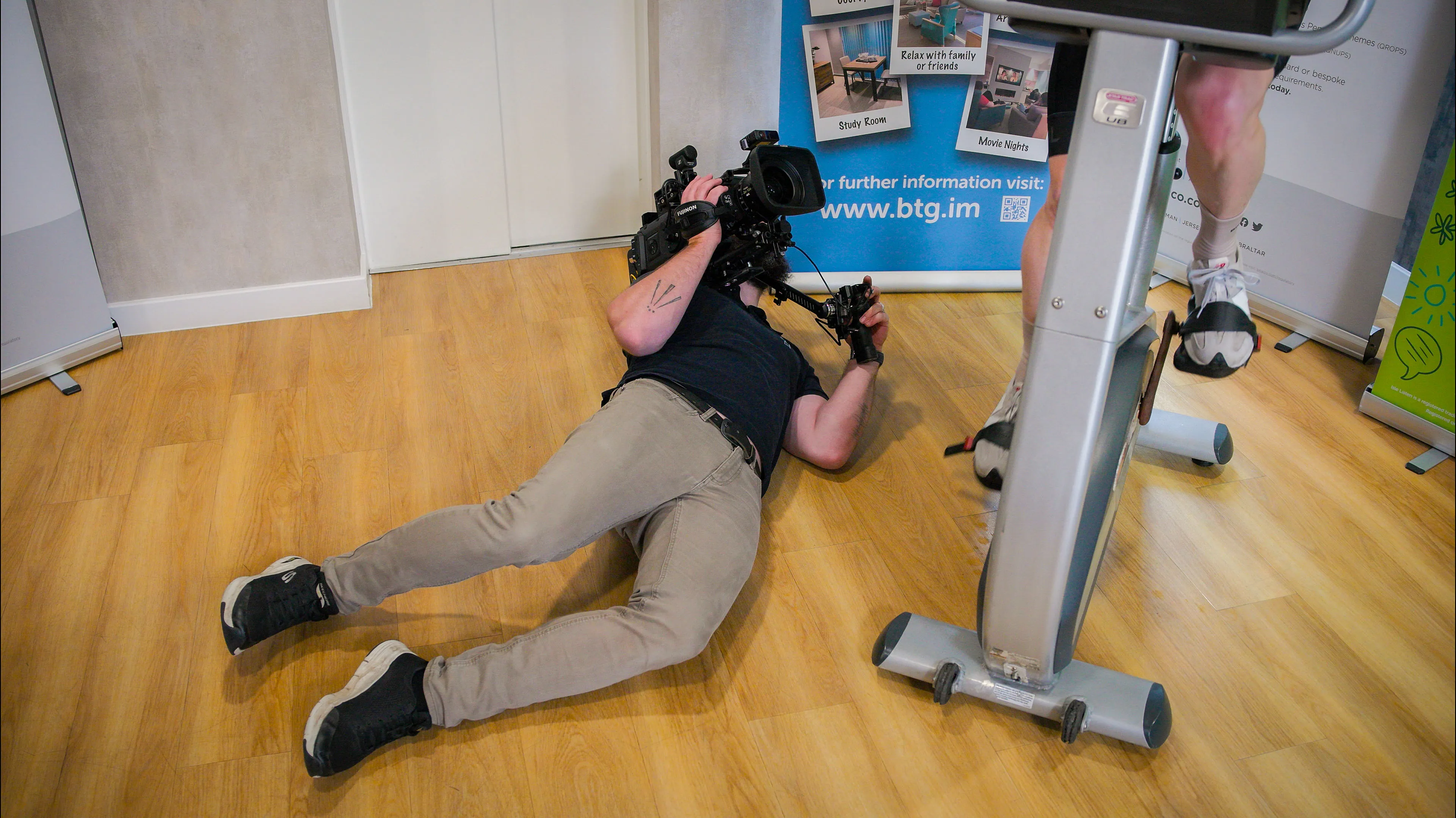 Nico van Loggerenberg from DotPerformance lying flat on the floor to shoot a low-angle shot with a Blackmagic URSA 12K Mini Pro beneath a static bike during the Boal & Co charity cycling challenge on the Isle of Man