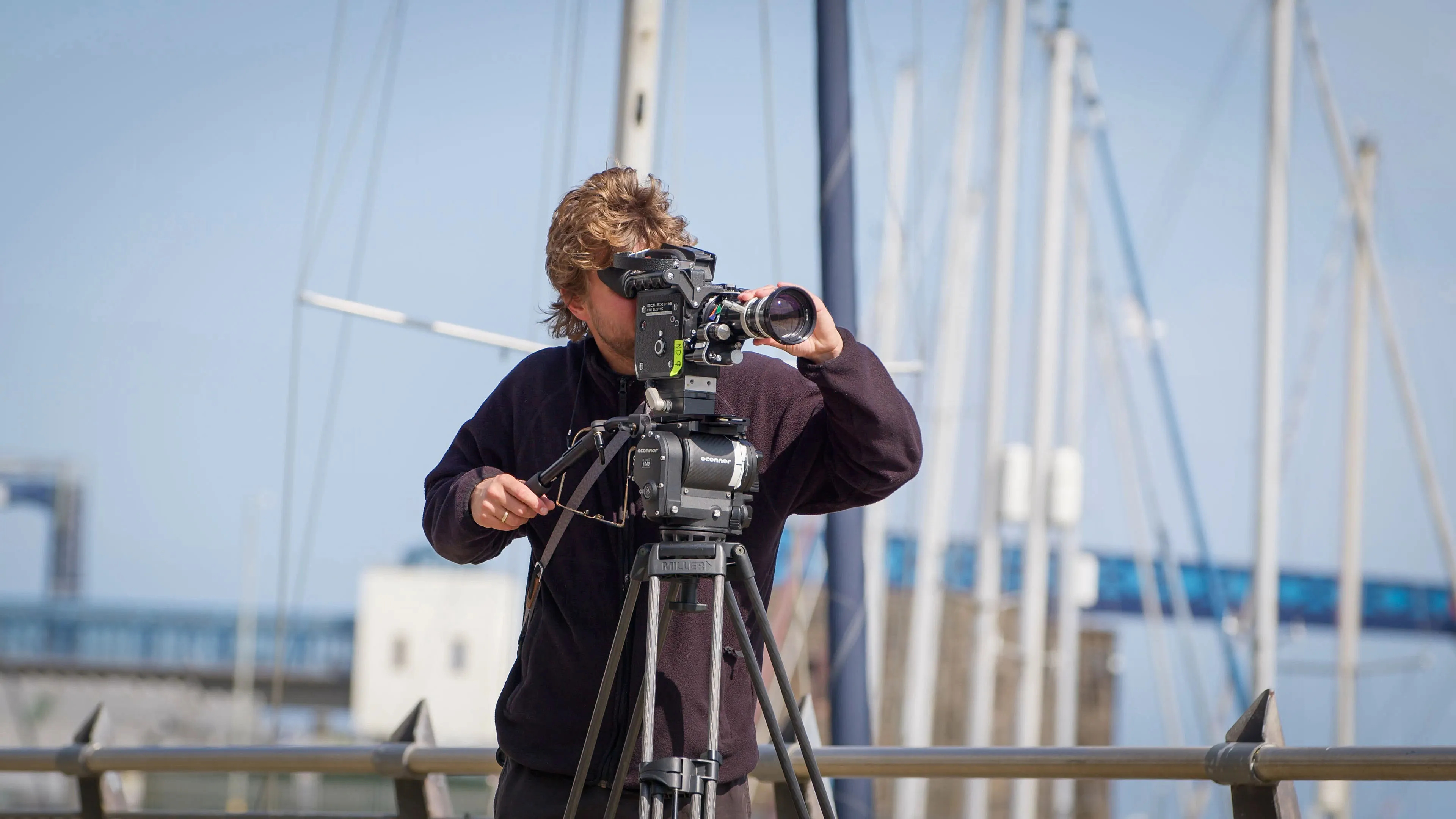A person operates a camera on a tripod at a marina, with blurred sailboat masts in the background. The scene conveys focus and technical skill.