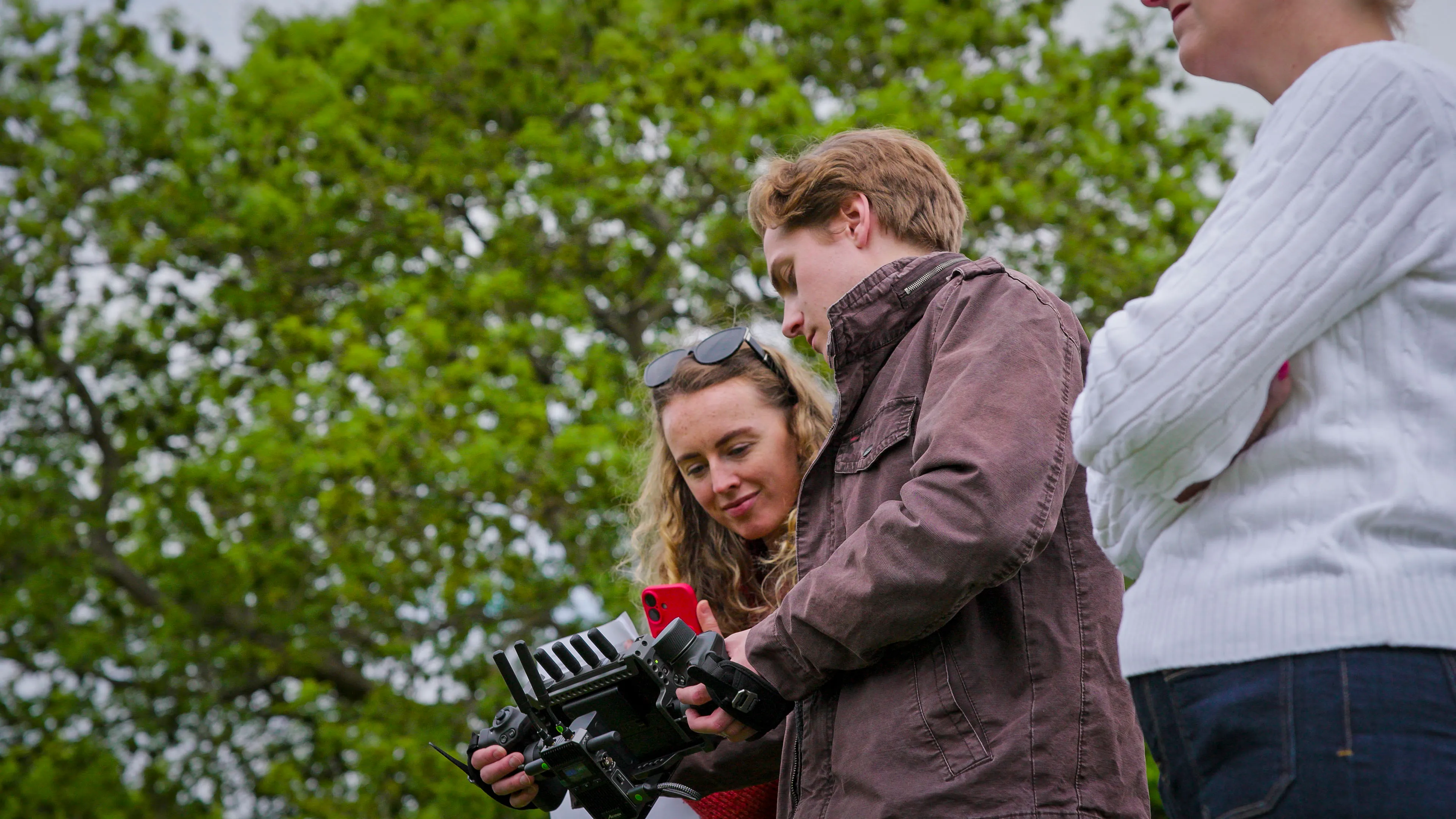 Three people outdoors examine a camera monitor. A woman in a red sweater and a man in a brown jacket focus on the device; another stands beside them. Green trees fill the backdrop, conveying a collaborative and curious atmosphere.