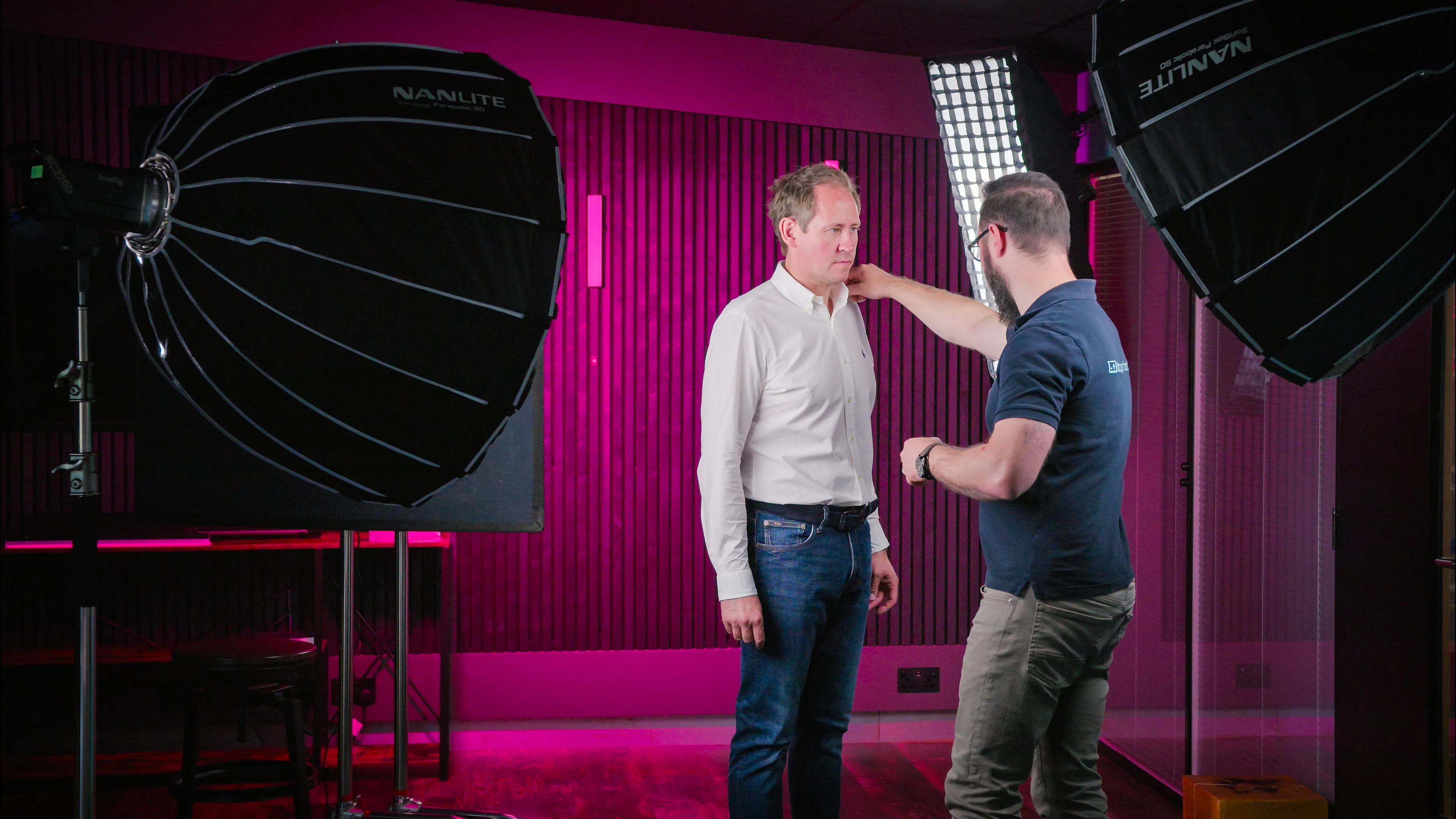A man stands as a photographer adjusts something in a studio with pink and purple hues. Large softbox lights create a professional setup. The atmosphere is focused and creative.